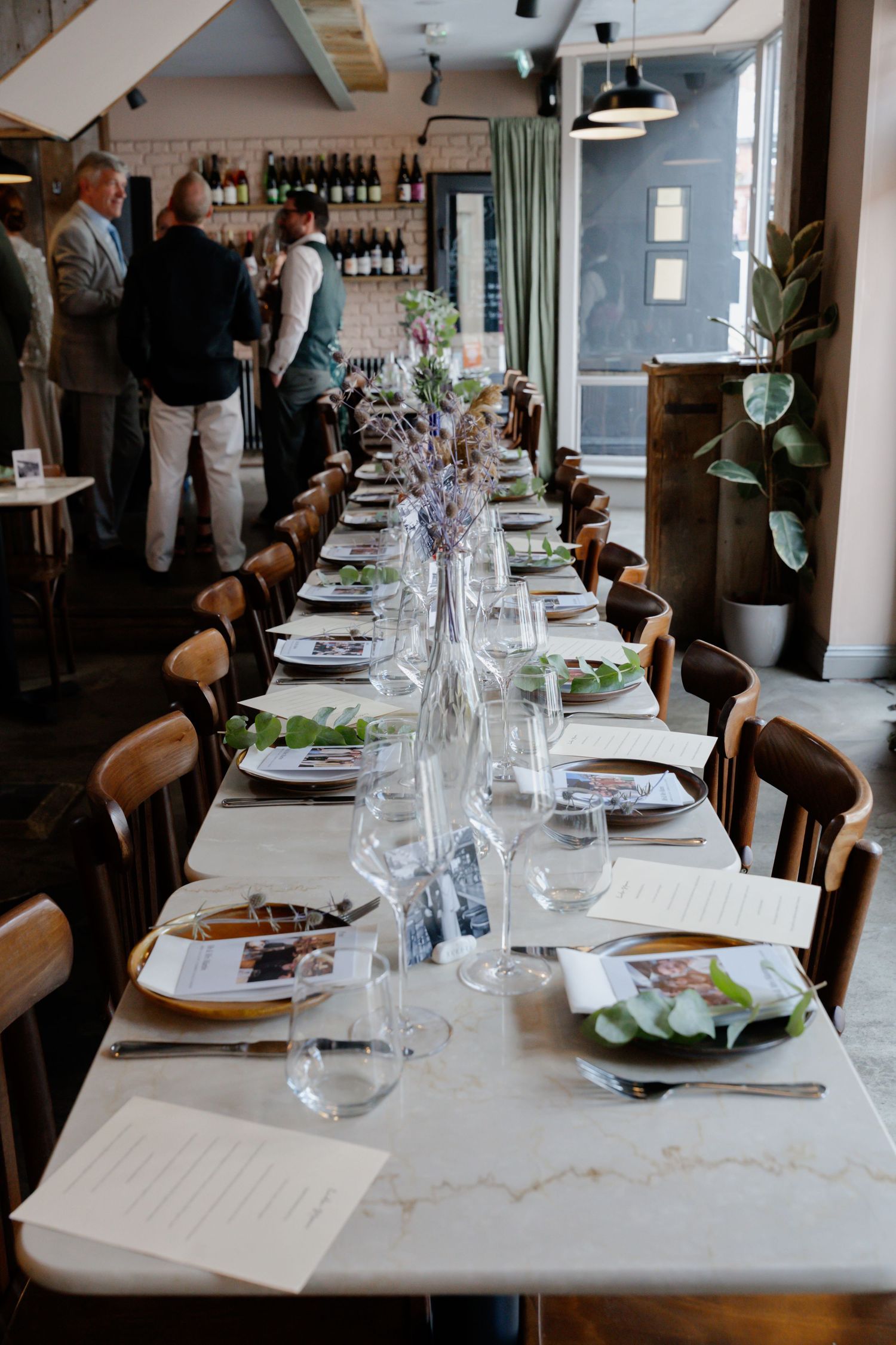 Elegant long wedding reception table set with glassware, menus, and floral centerpieces in a restaurant.
