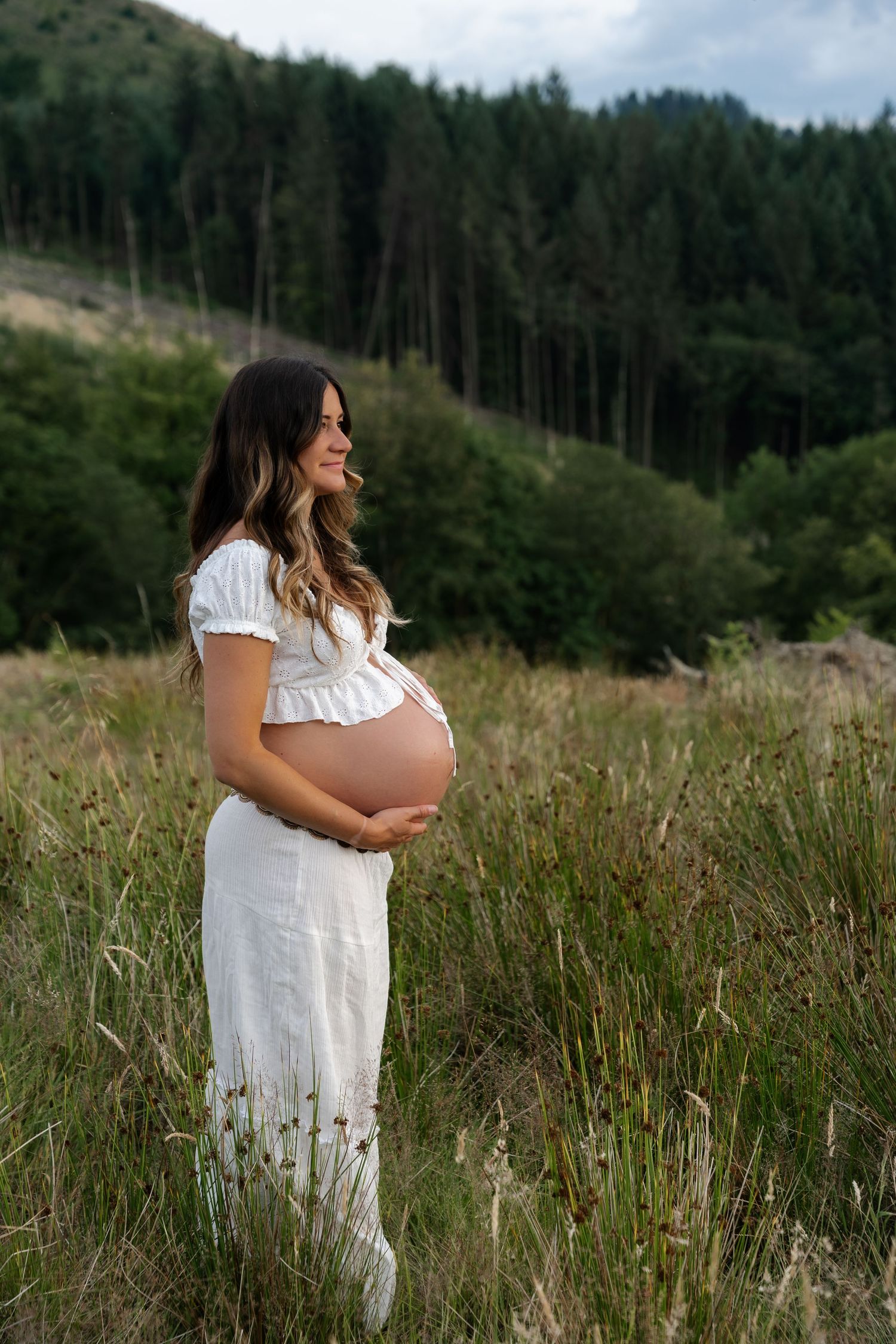 Smiling pregnant woman in white dress holding belly in a green field.