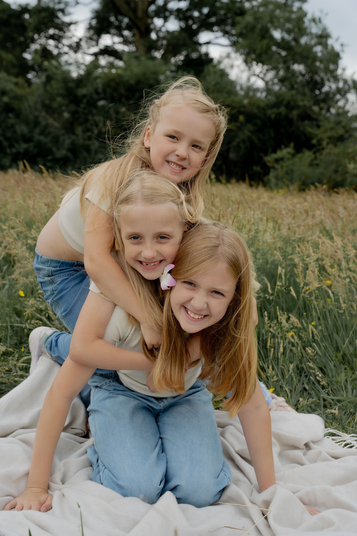 Three blonde sisters playfully stacking on each other on blanket in green field, smiling.