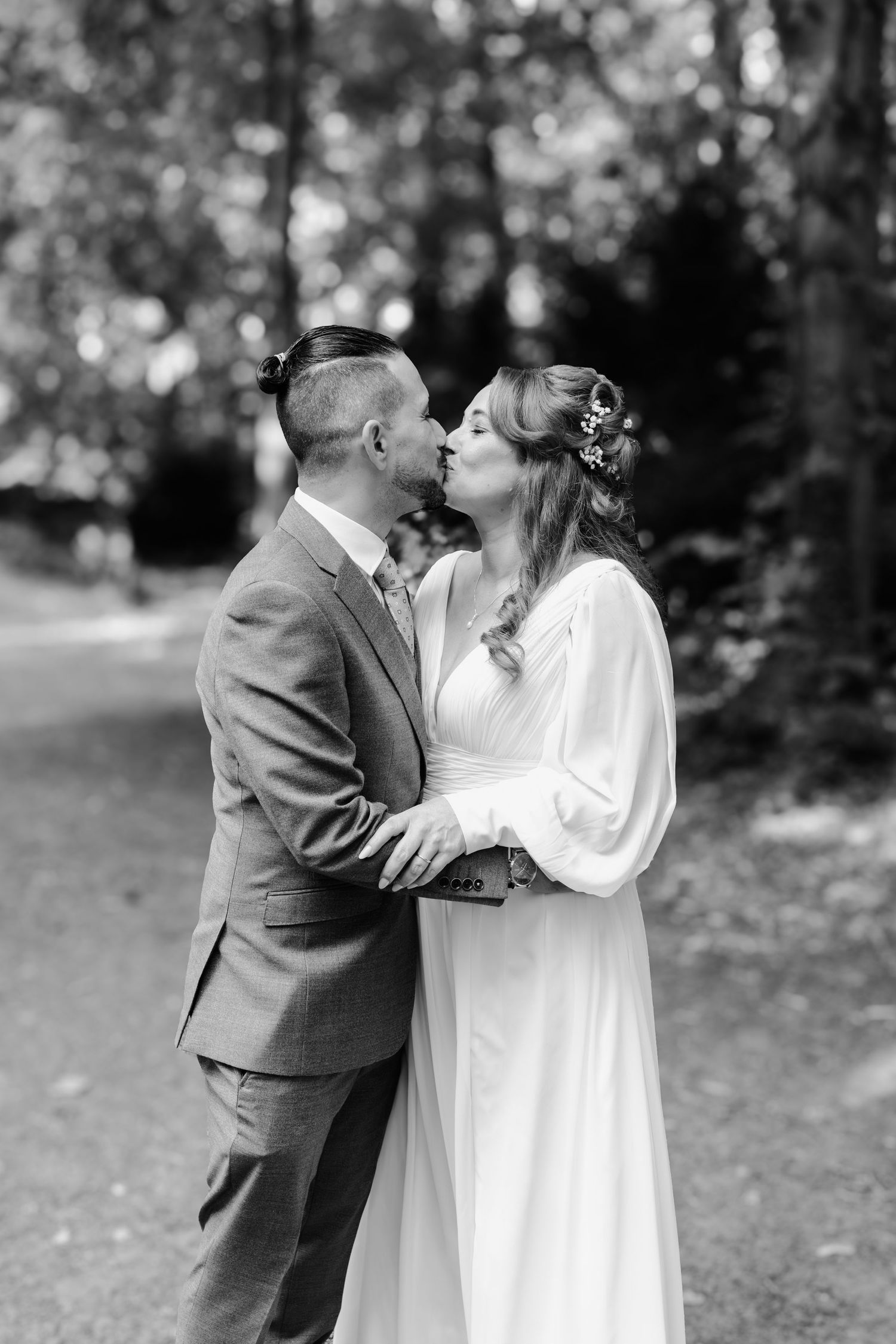 Bride and groom sharing romantic kiss on tree-lined path in elegant black and white wedding portrait.