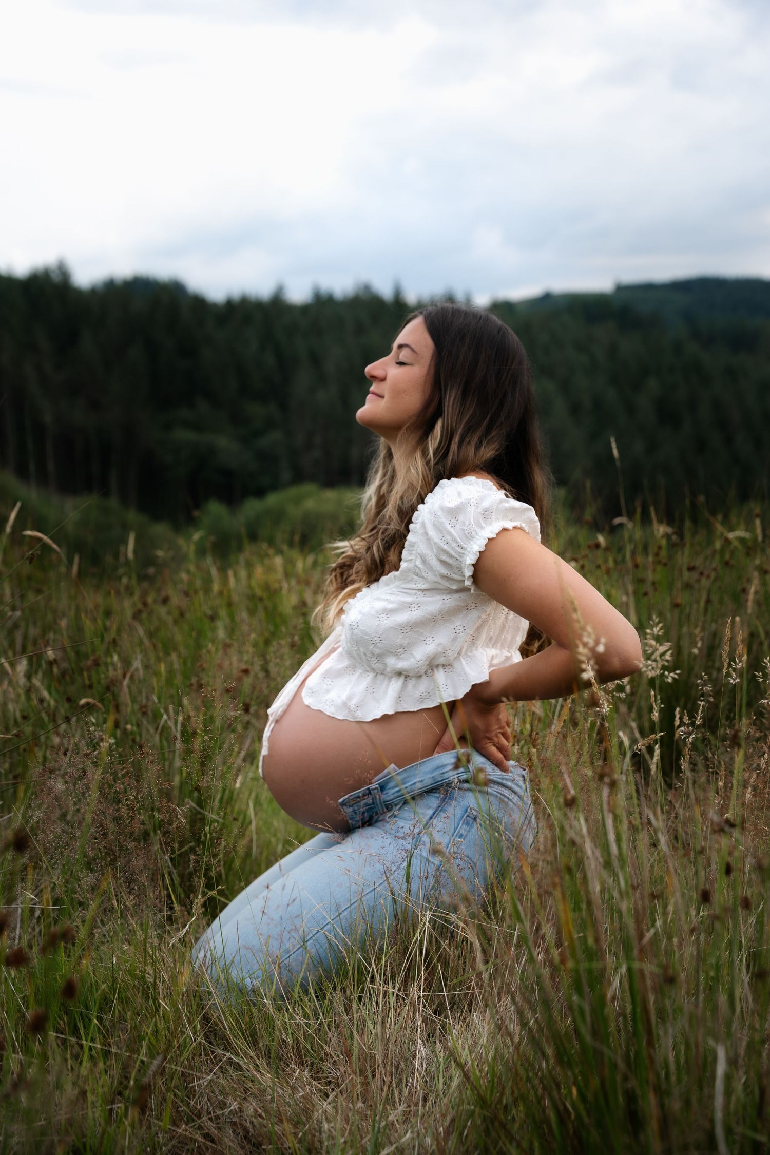 Pregnant woman in white crop top and jeans kneeling in a grassy field.