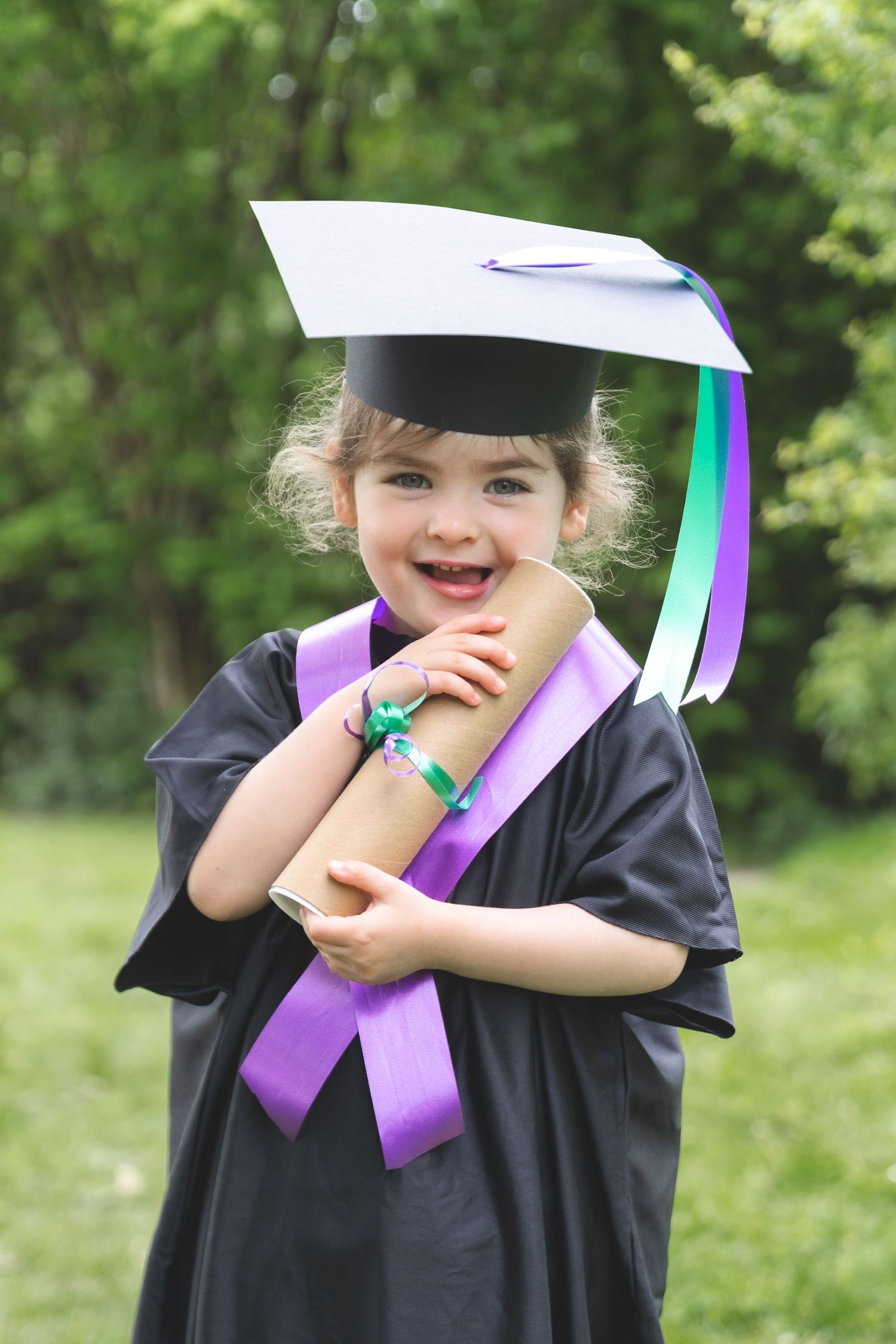 A cheerful little girl wearing a black graduation gown and mortarboard hat holds a diploma scroll outdoors.