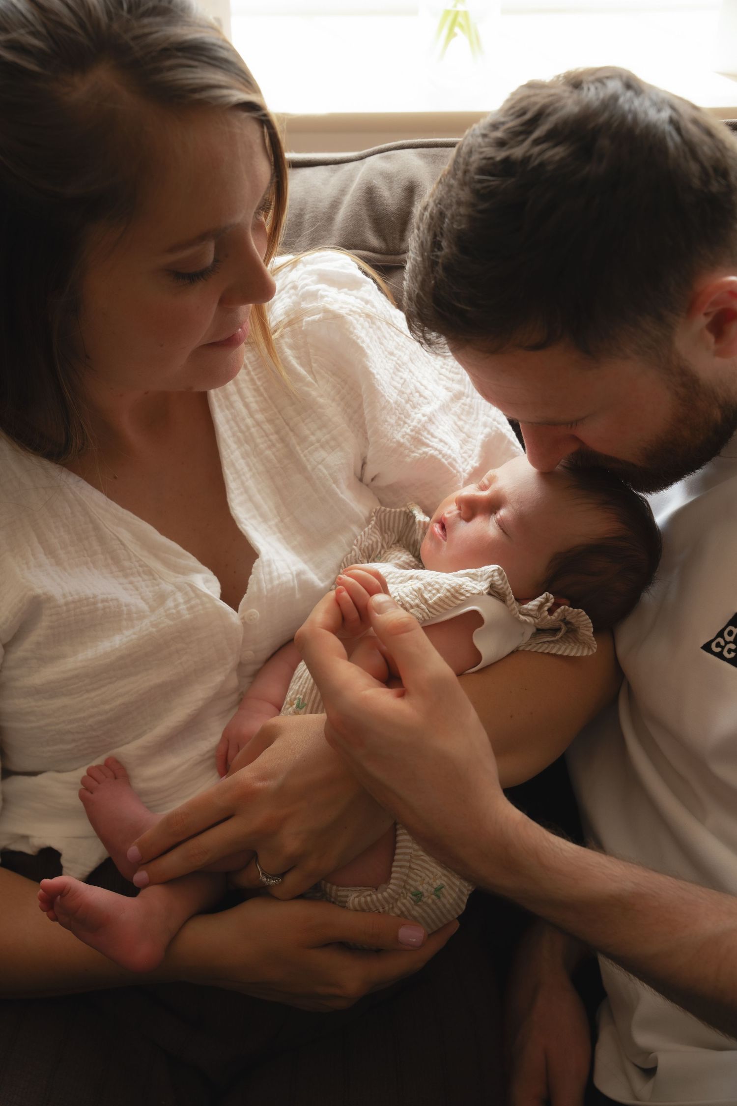 New parents tenderly hold and kiss their sleeping newborn baby together on a couch in a warm home setting.