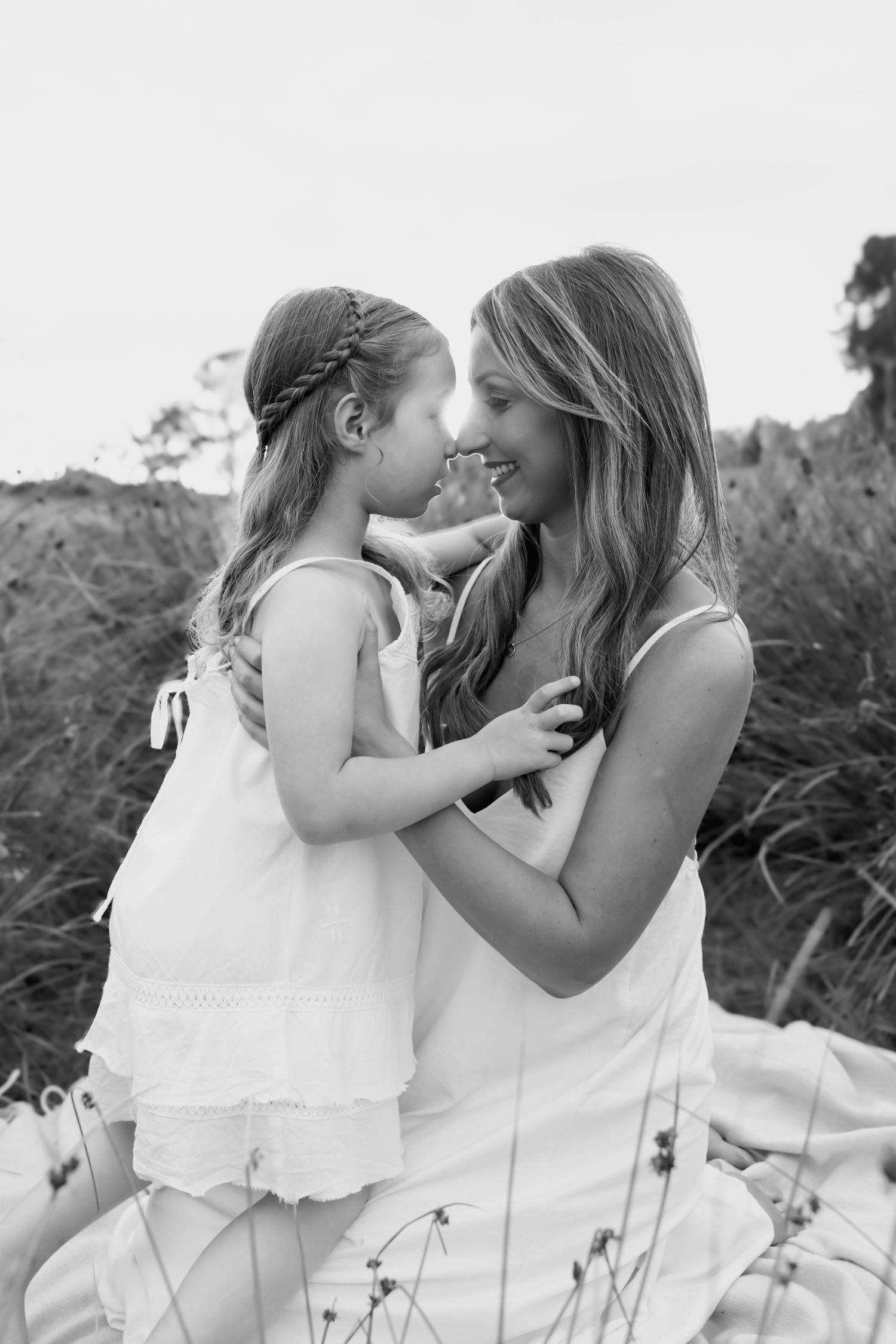 Black and white photo of a smiling mother and daughter touching noses in a field.