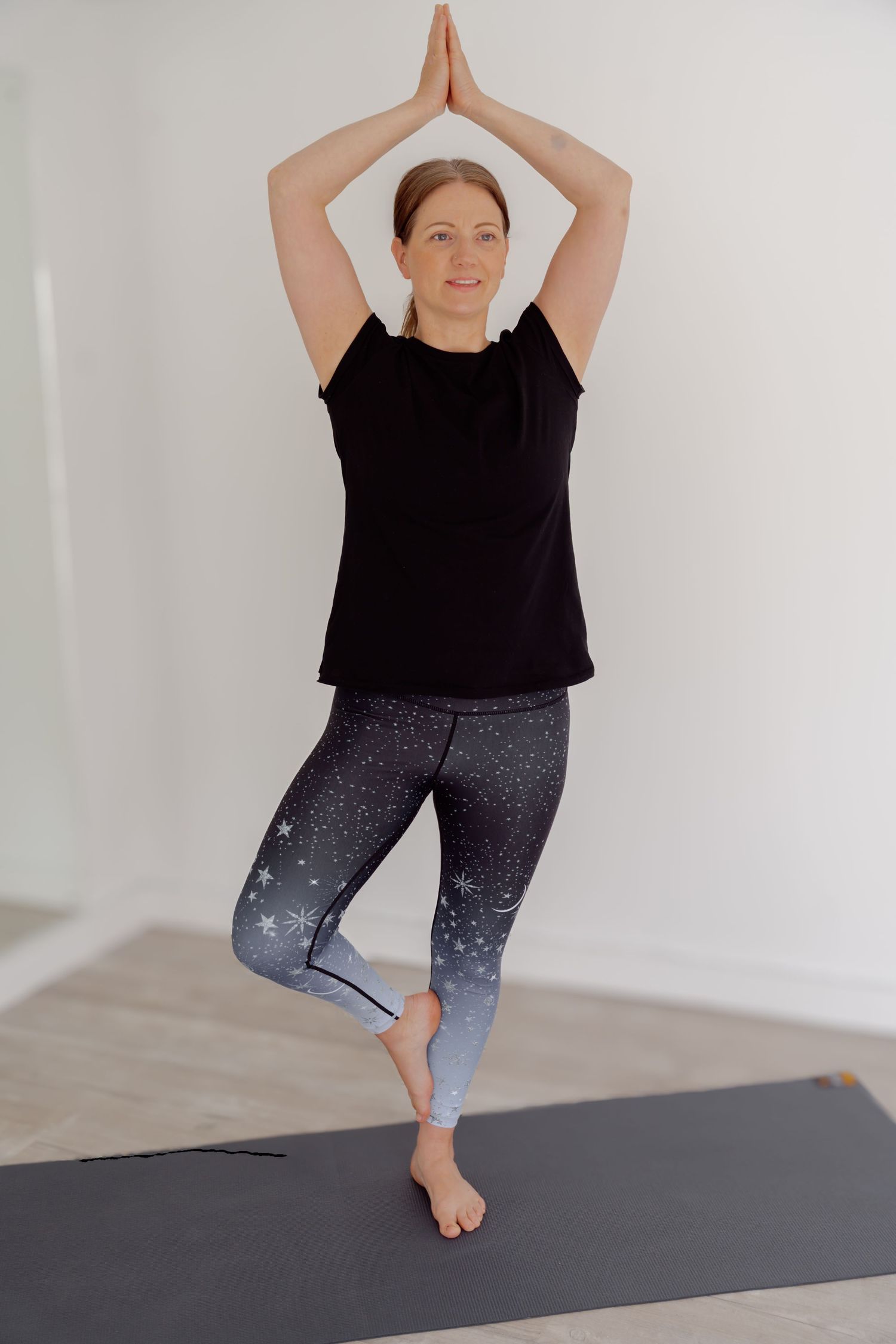 Woman in black top and galaxy print leggings performing tree pose on yoga mat in bright white studio.