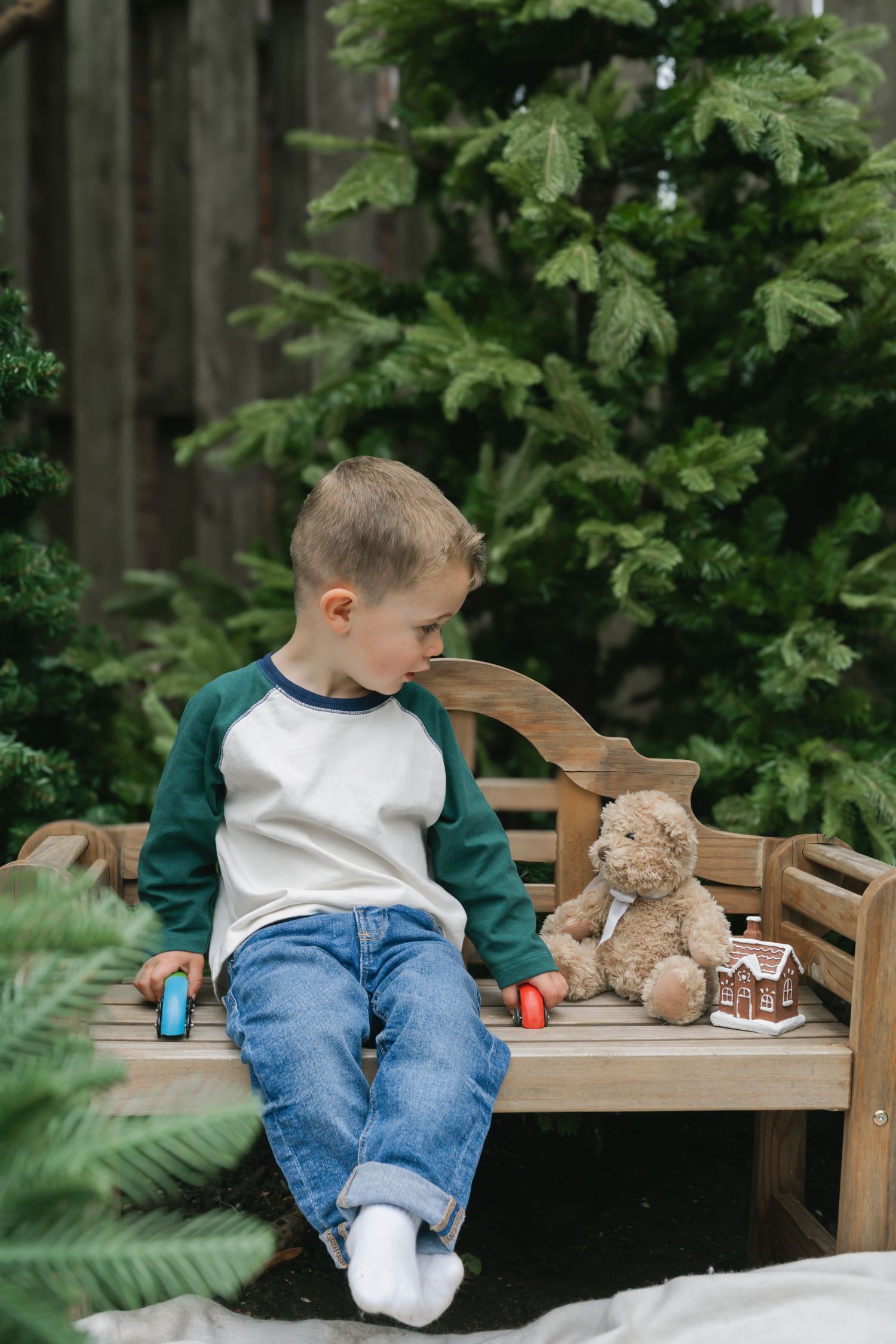 Young boy in jeans sits on wooden bench with teddy bear and toy house near evergreen trees outdoors.