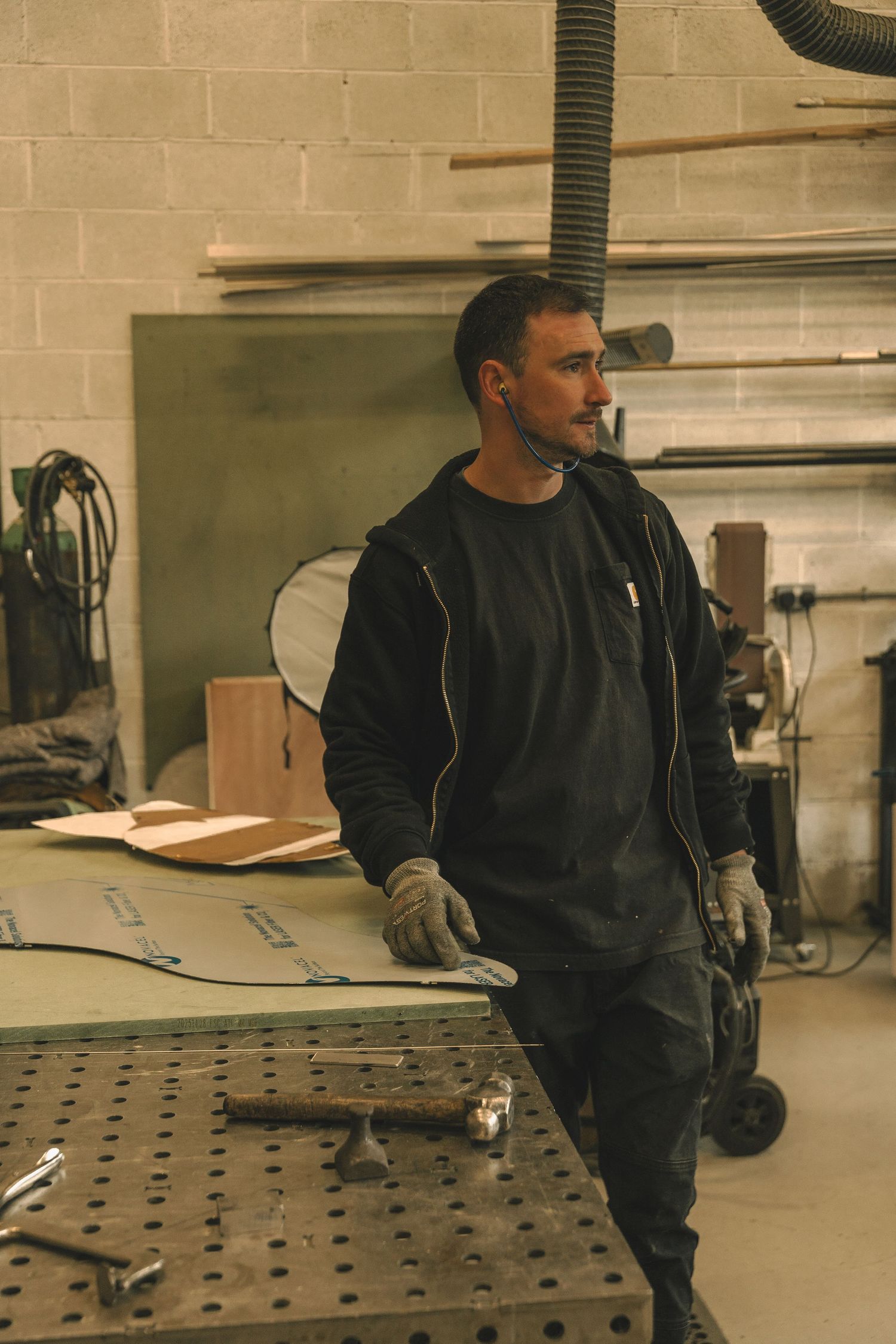 A male metalworker in black workwear and gloves stands at a workshop table surrounded by tools and materials.