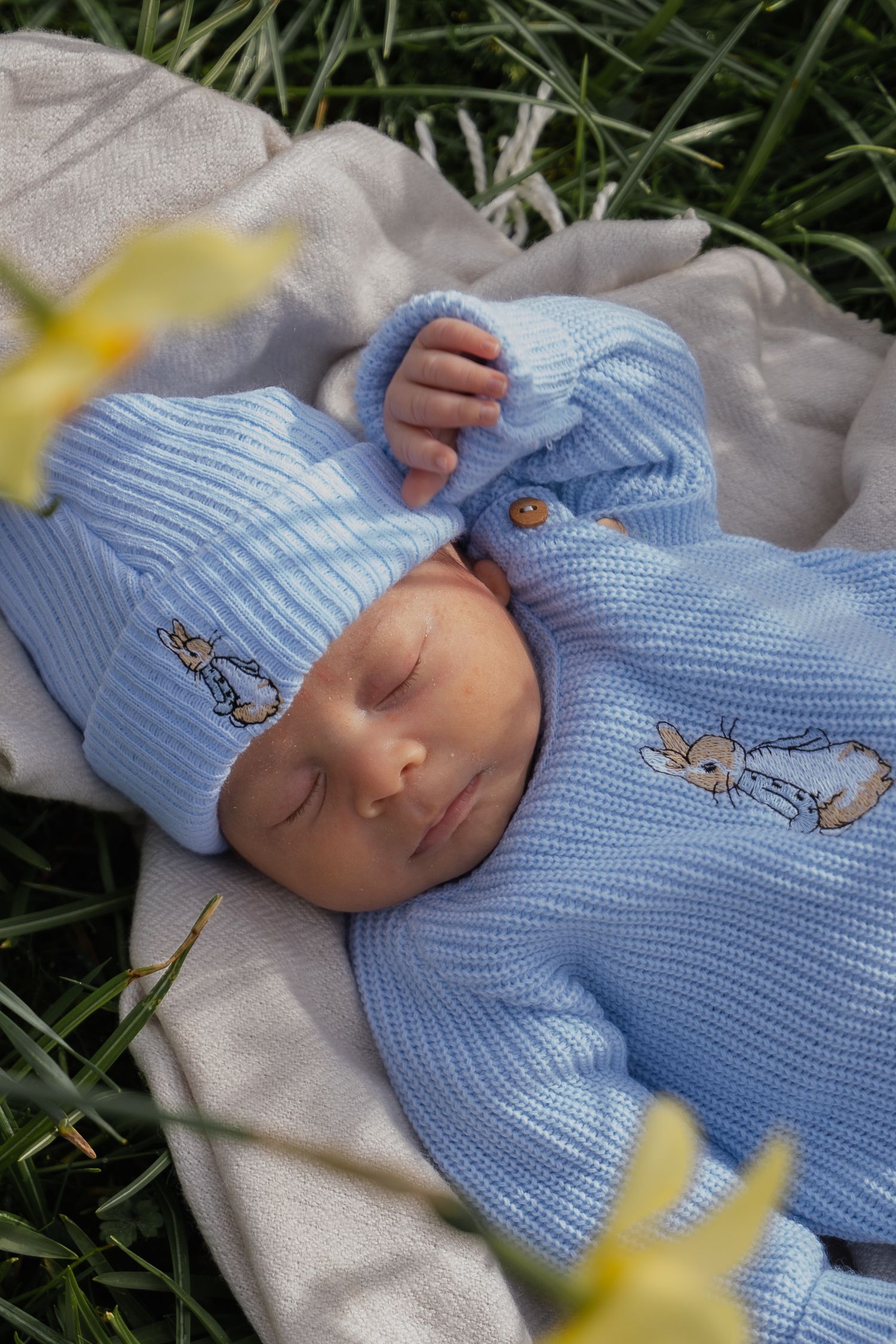Sleeping newborn in blue Peter Rabbit knit outfit and hat lying on a blanket surrounded by green grass and daffodils.