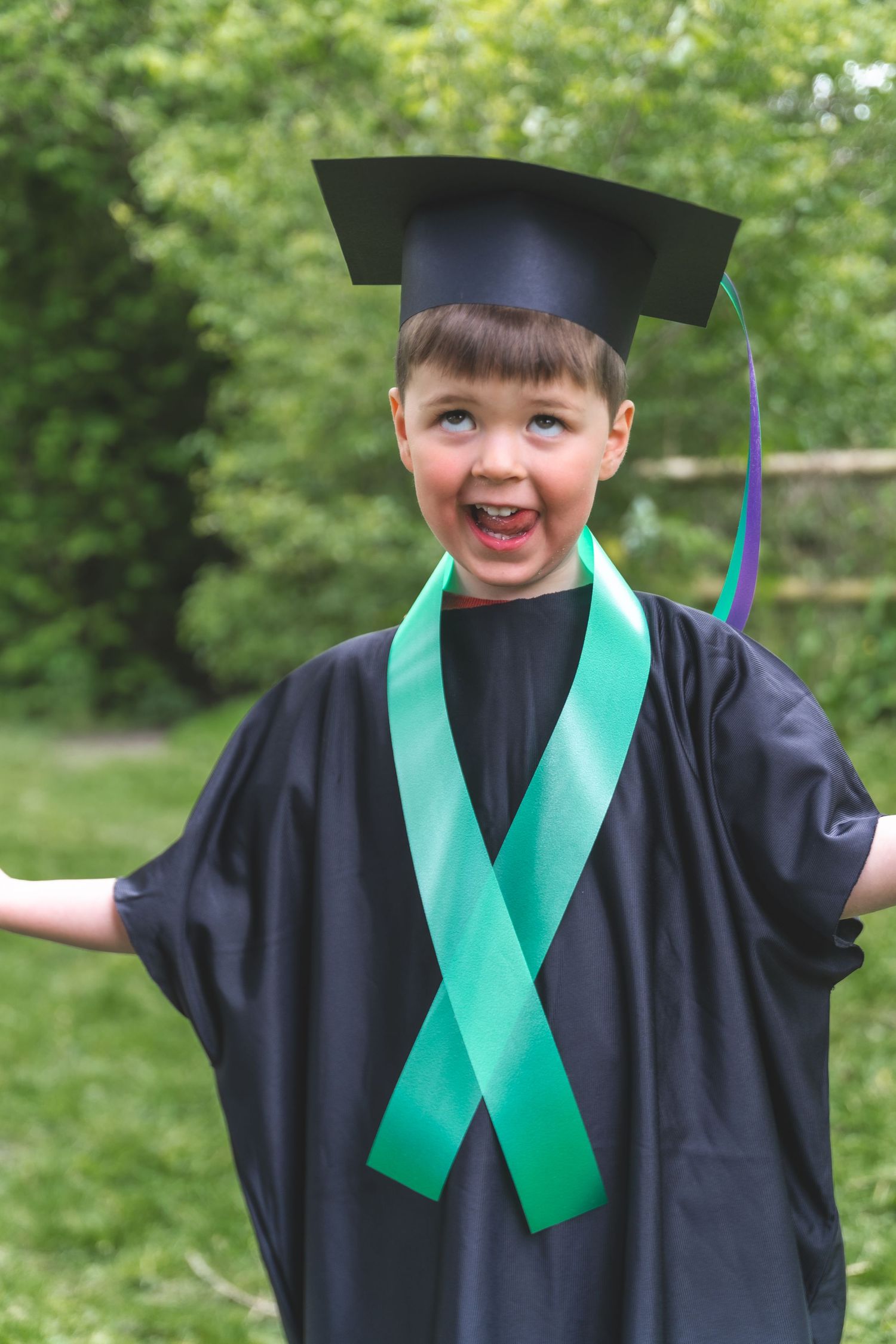 Happy young boy in black graduation gown and cap with green sash, smiling outdoors.