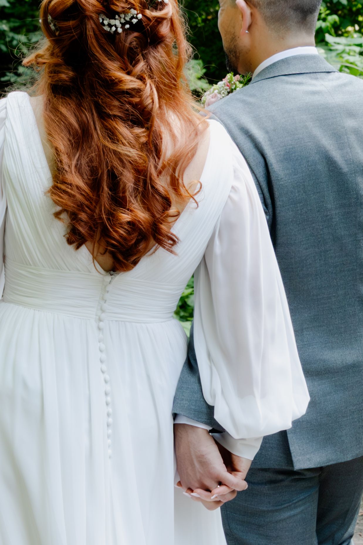 Redhead bride with floral hair accessories holding hands with groom, showing back of white wedding dress.