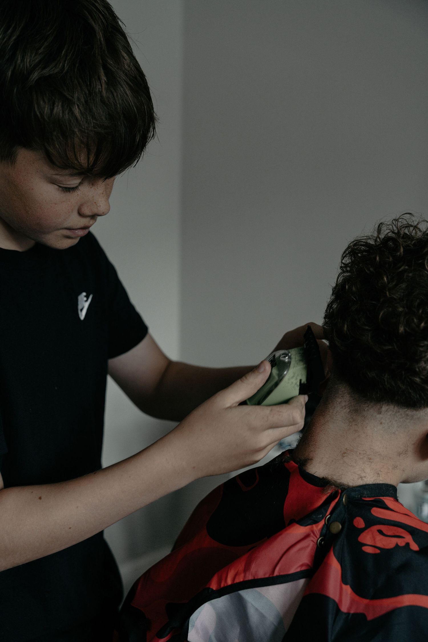 A focused young boy in a black t-shirt uses green hair clippers to cut another childs curly hair.