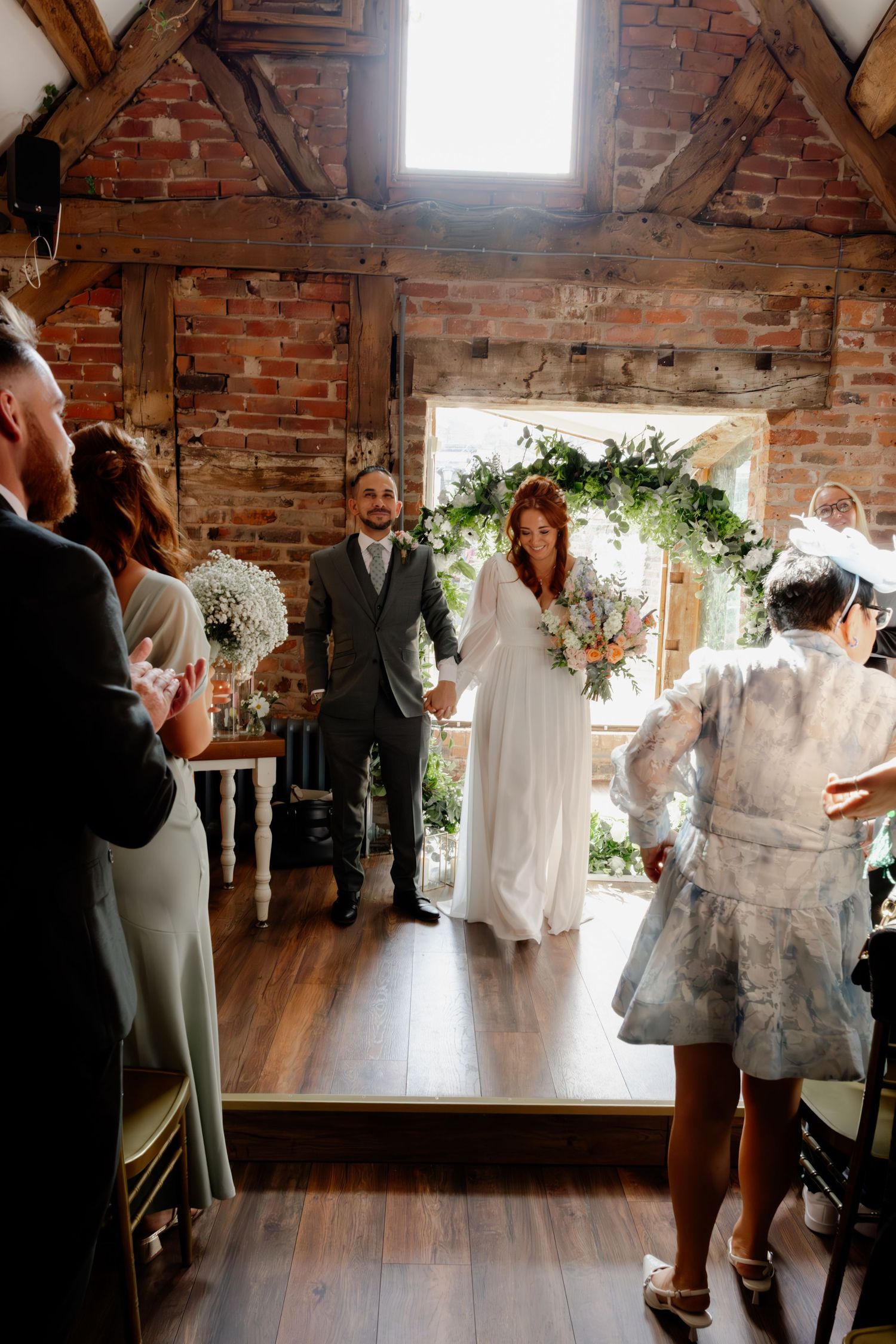 Bride and groom standing at floral arch altar in rustic brick barn ceremony venue.