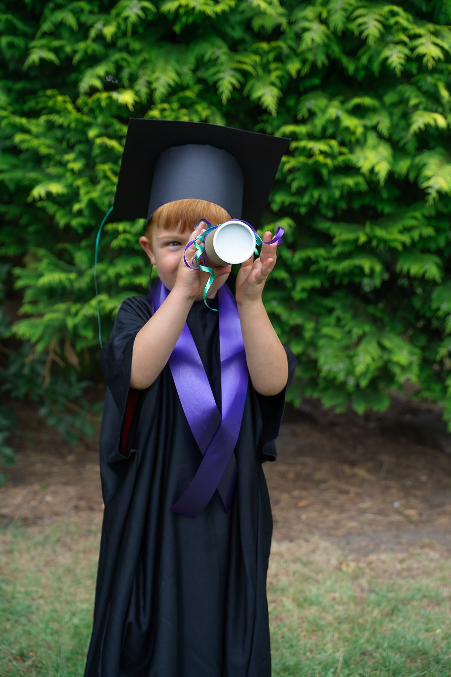 Red-haired child in graduation gown and cap looking through rolled diploma outdoors.
