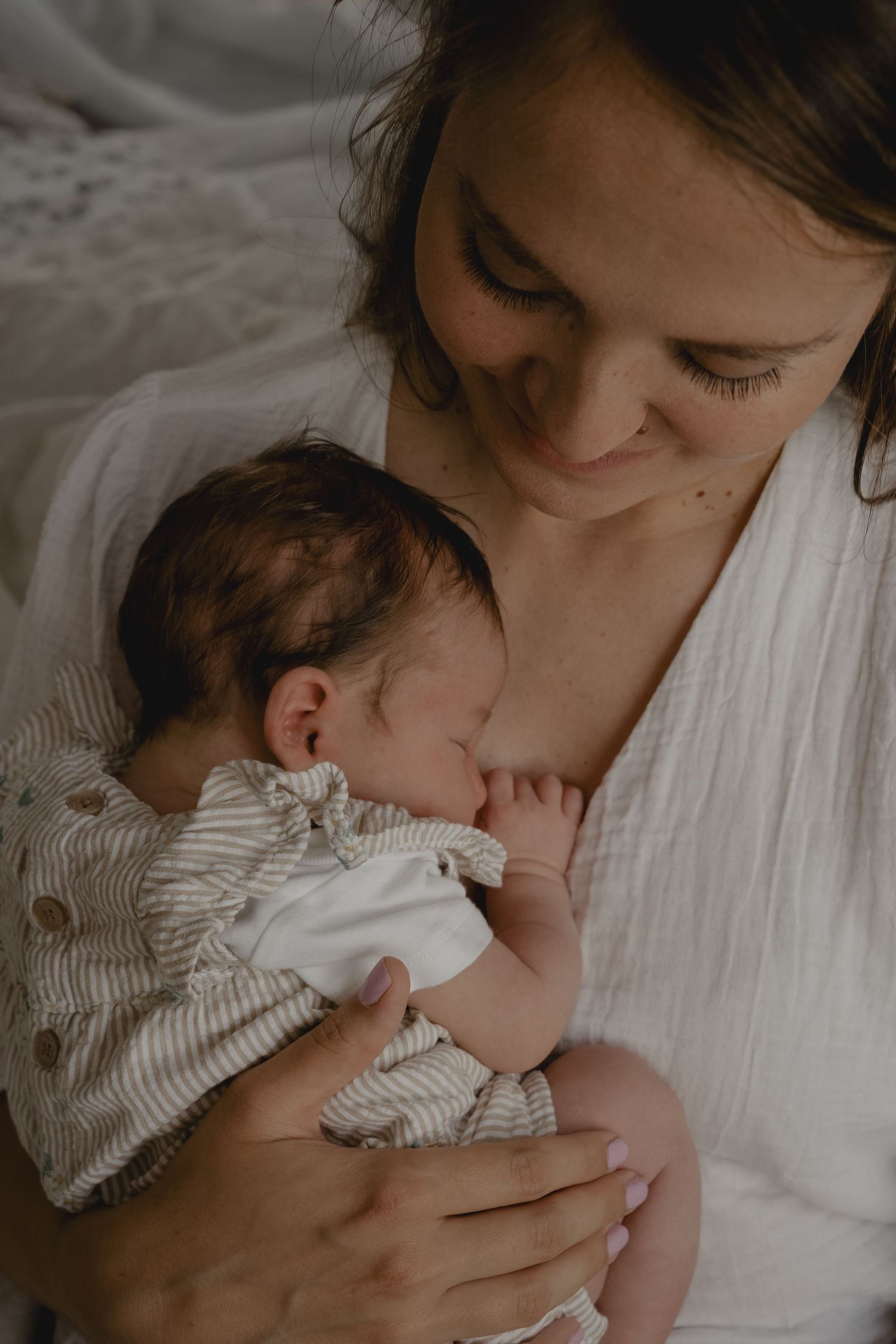 Mother in white robe tenderly breastfeeding her newborn baby dressed in striped onesie.