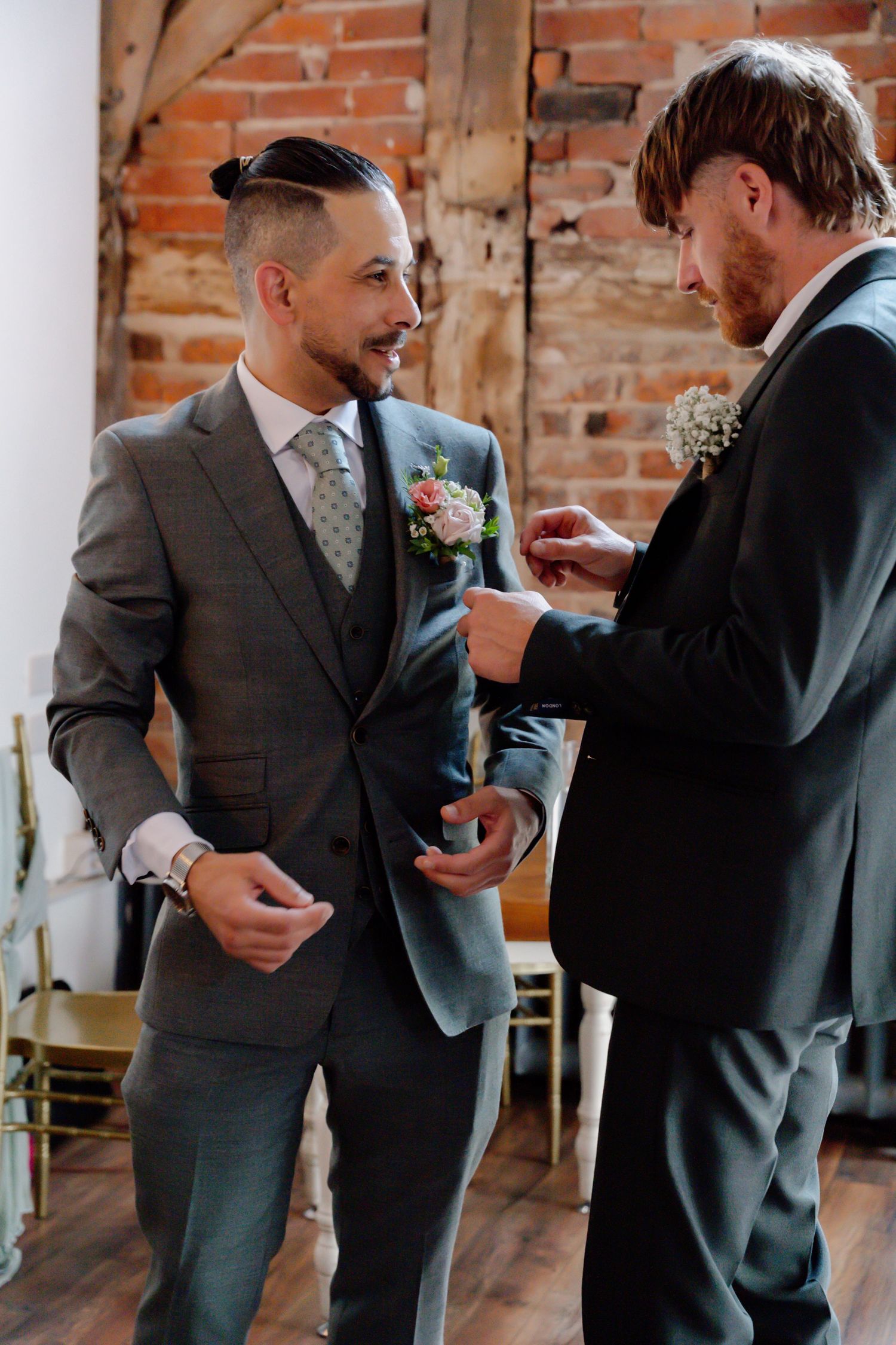 Two men in gray suits at wedding venue, one pinning boutonniere on the other.
