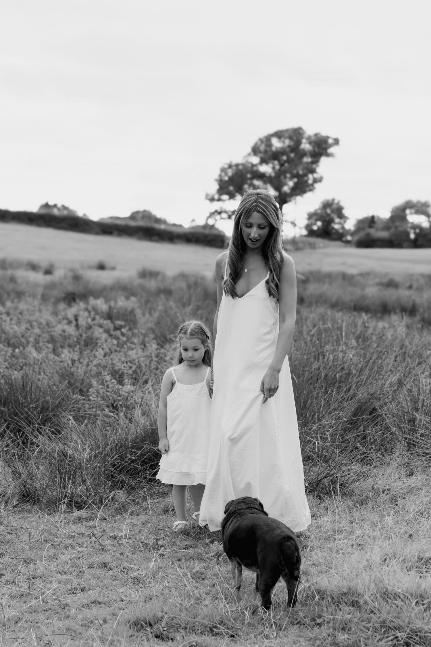 Black and white photo of mother, daughter, and dog walking in a countryside field.