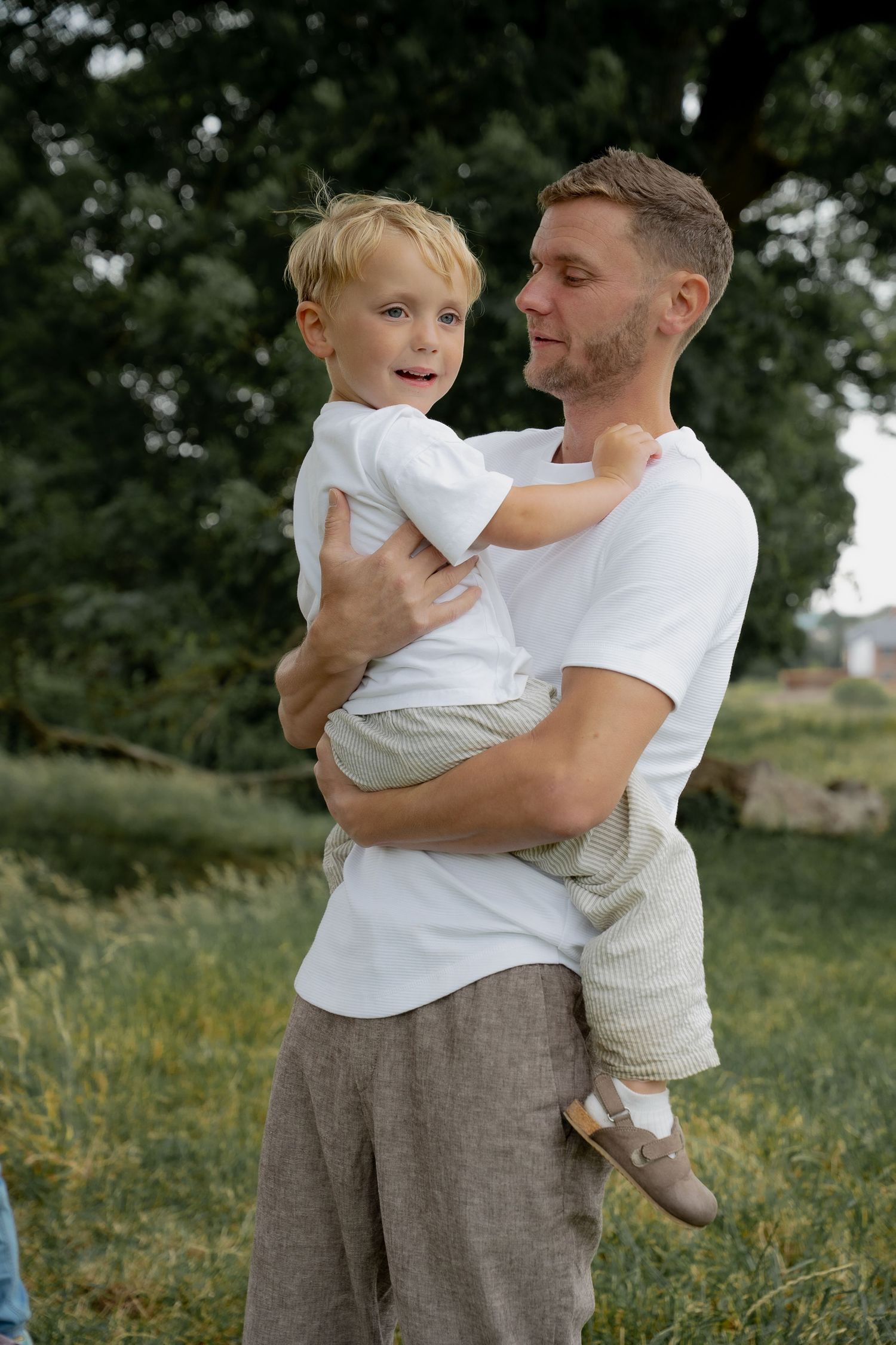 Smiling father holds young blonde toddler son in arms outdoors in green nature setting.