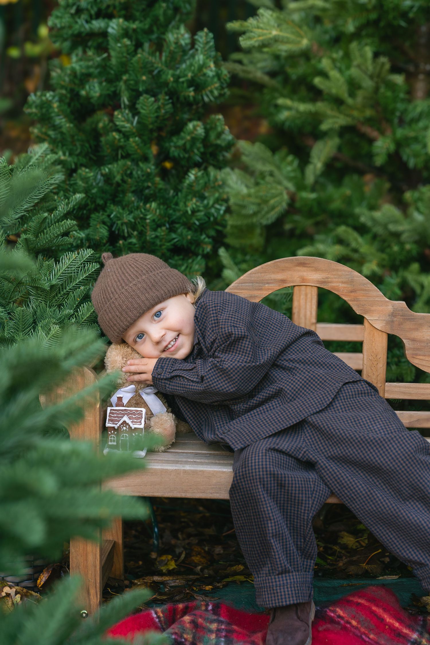 Smiling child in brown hat and gray suit lounges on wooden bench surrounded by green pine trees with gingerbread house toy.