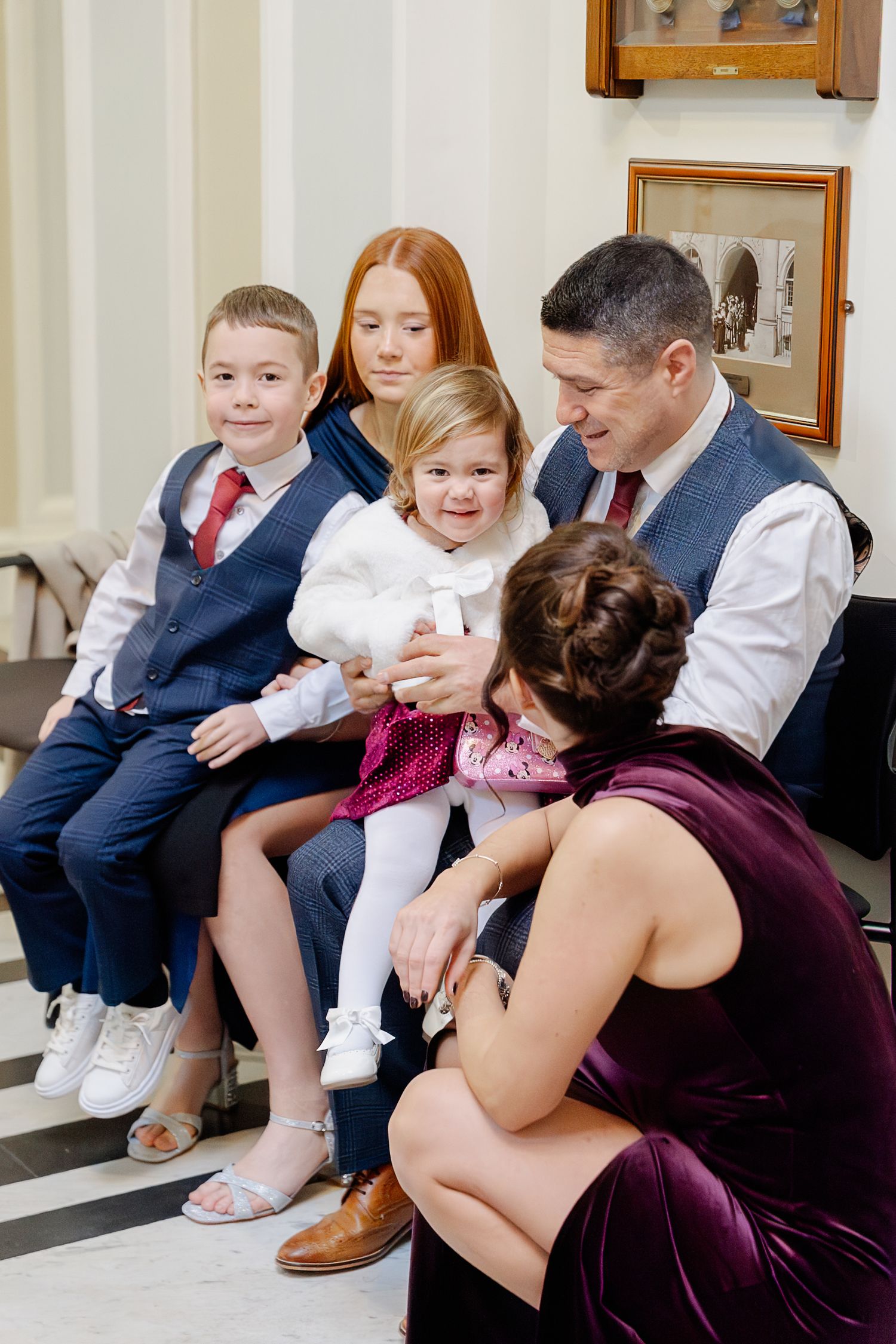 Family gathering with parents, two children, and a woman in purple velvet dress admiring a smiling toddler girl in white.