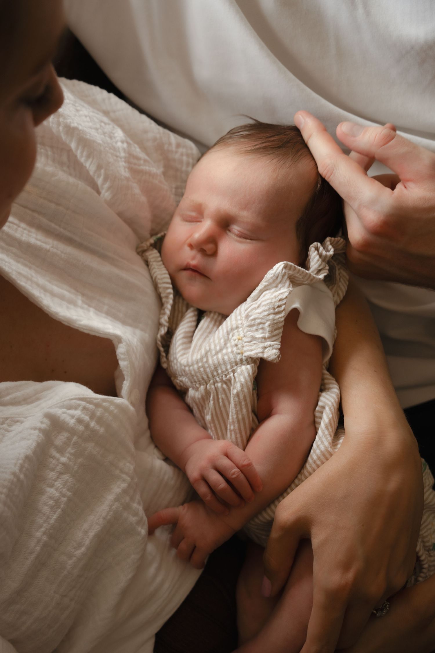Sleeping newborn baby in white textured romper cradled in parents arms, peaceful infant portrait.