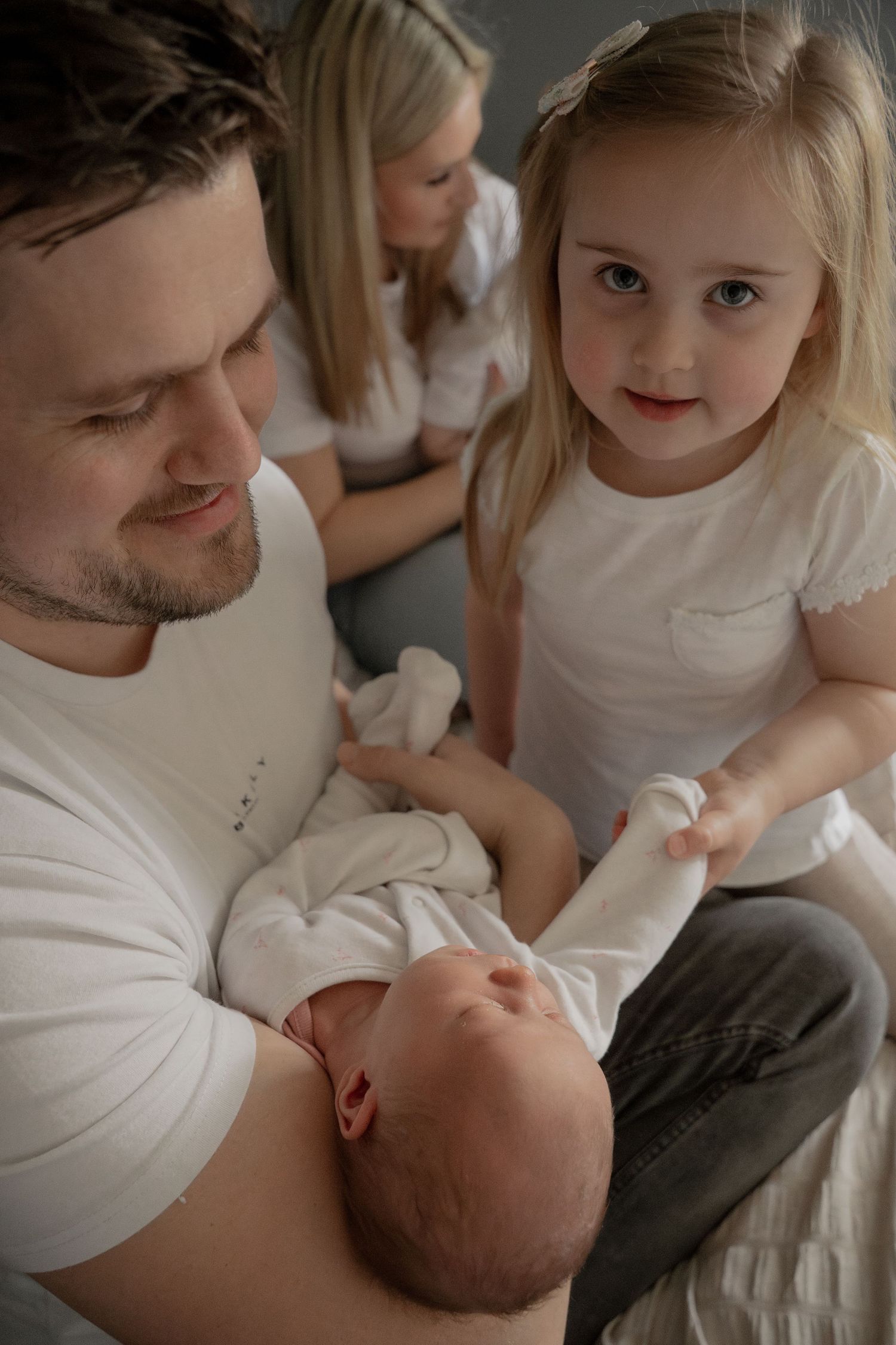Father holds newborn baby while young blonde girl touches infant's hand during family bonding moment.