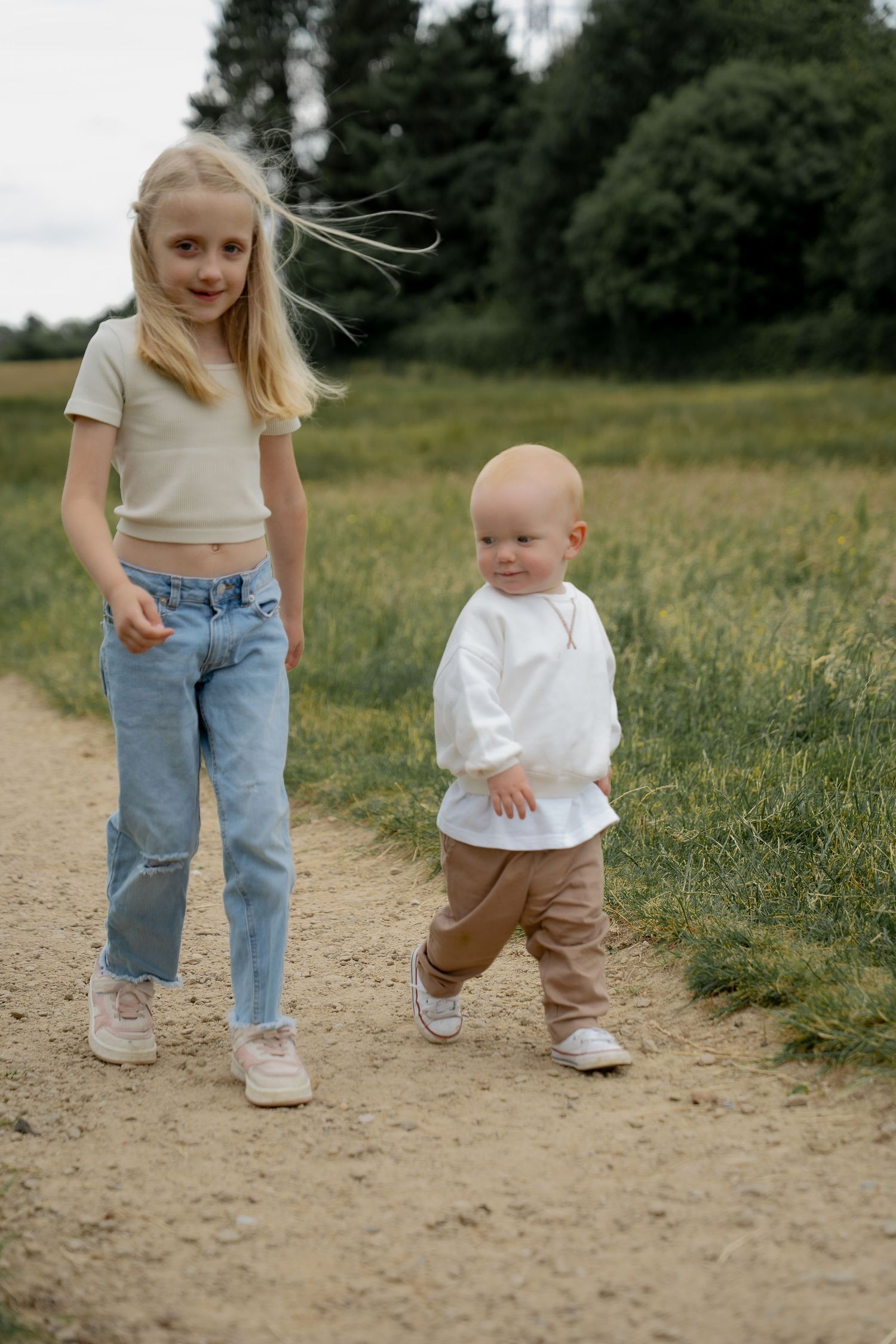 Young blonde girl and toddler sibling walking together on dirt path in countryside field.