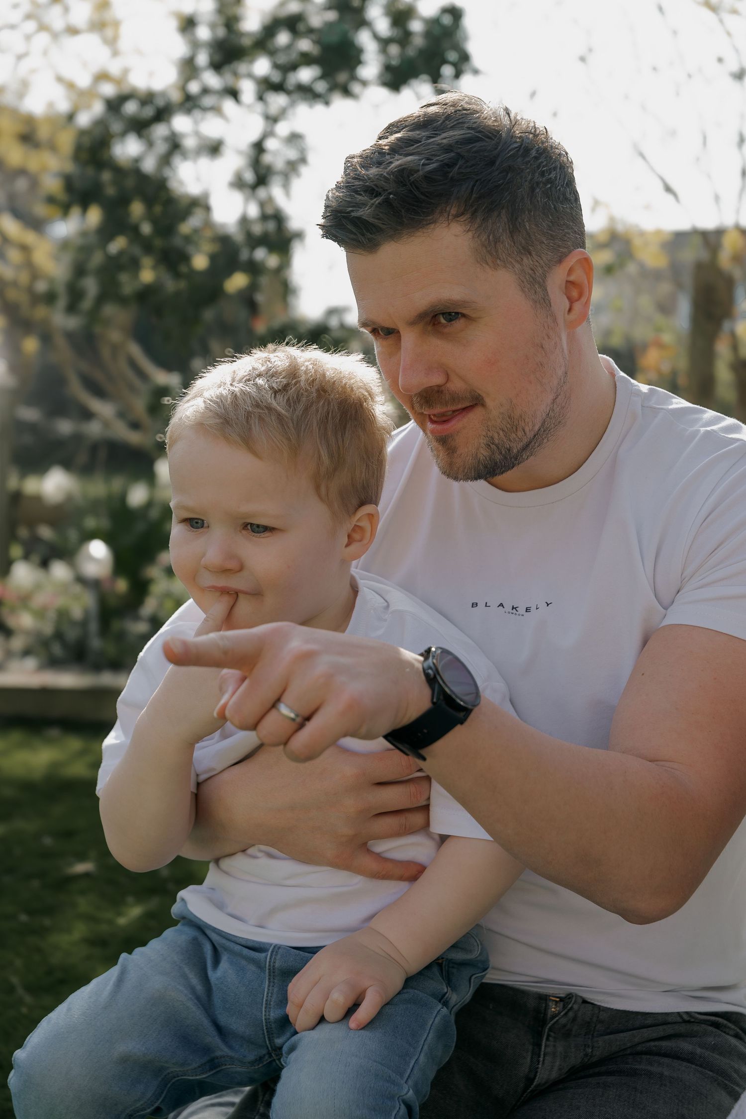Father in white t-shirt sitting outdoors with his young blonde toddler son on a sunny garden day.