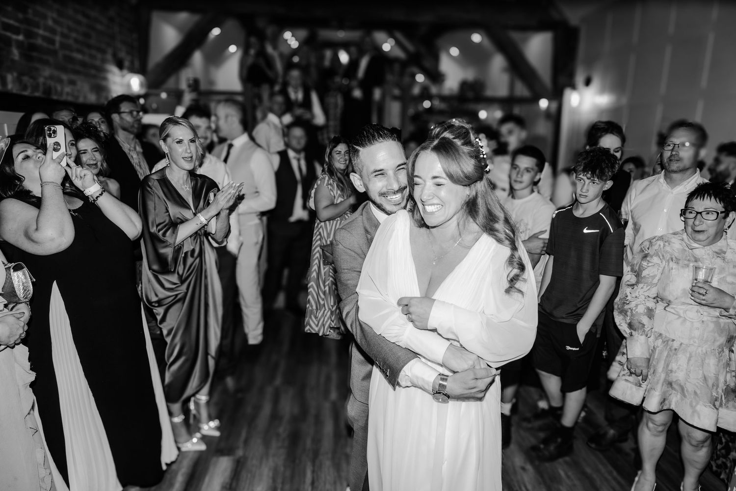 Black and white photo of bride and groom sharing first dance surrounded by cheering wedding guests.