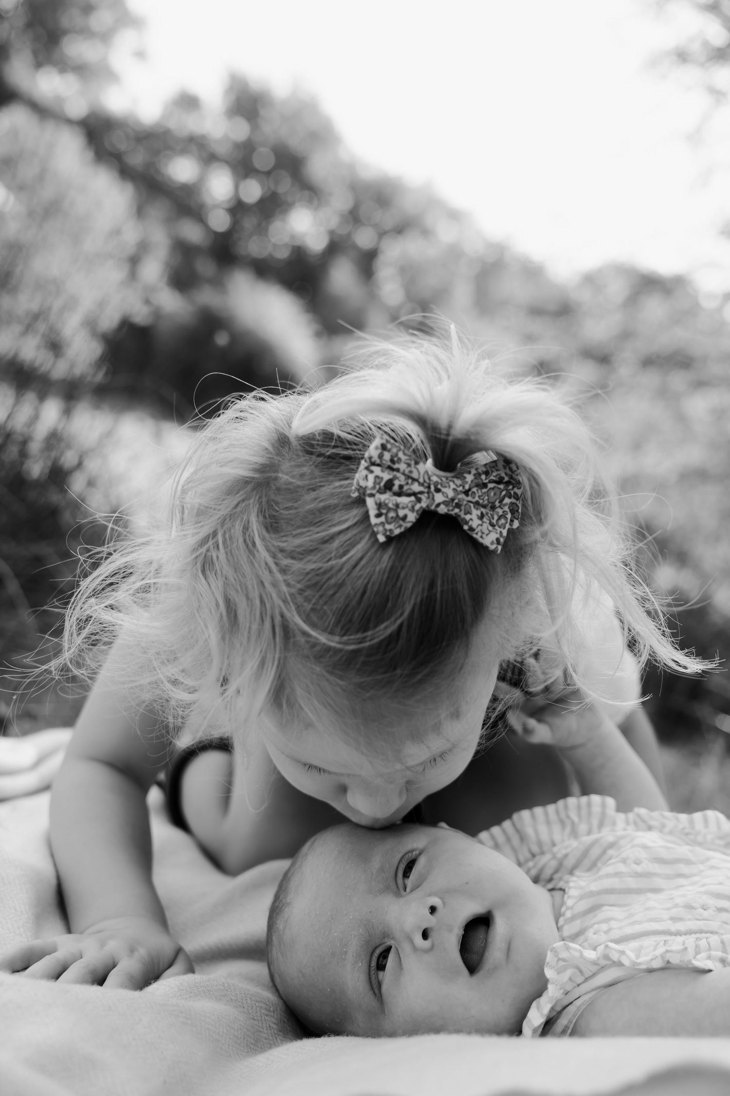 Black and white photo of young girl gently kissing her newborn sibling outdoors.