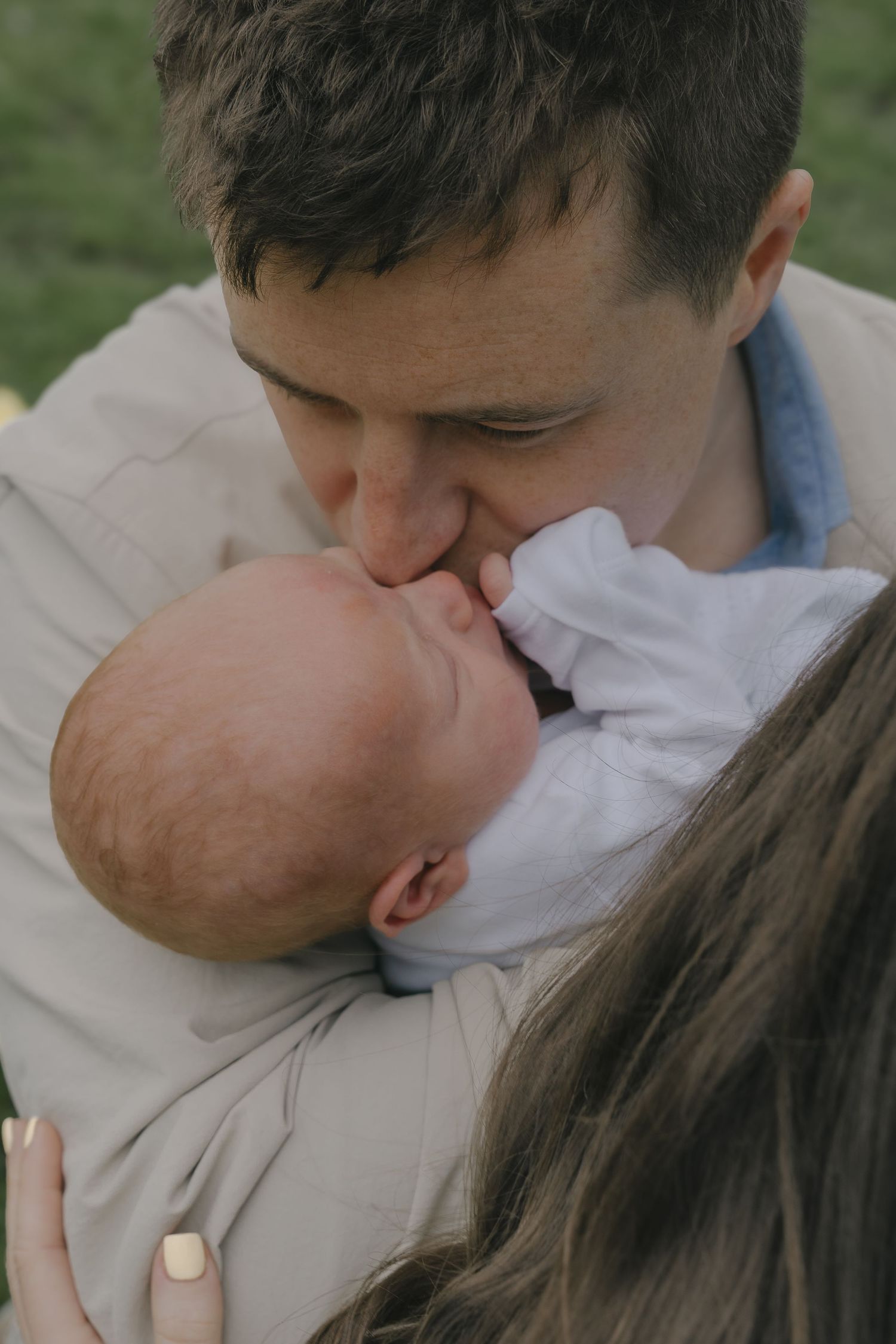 A father tenderly kissing his newborn baby while holding them close outdoors on a green lawn.