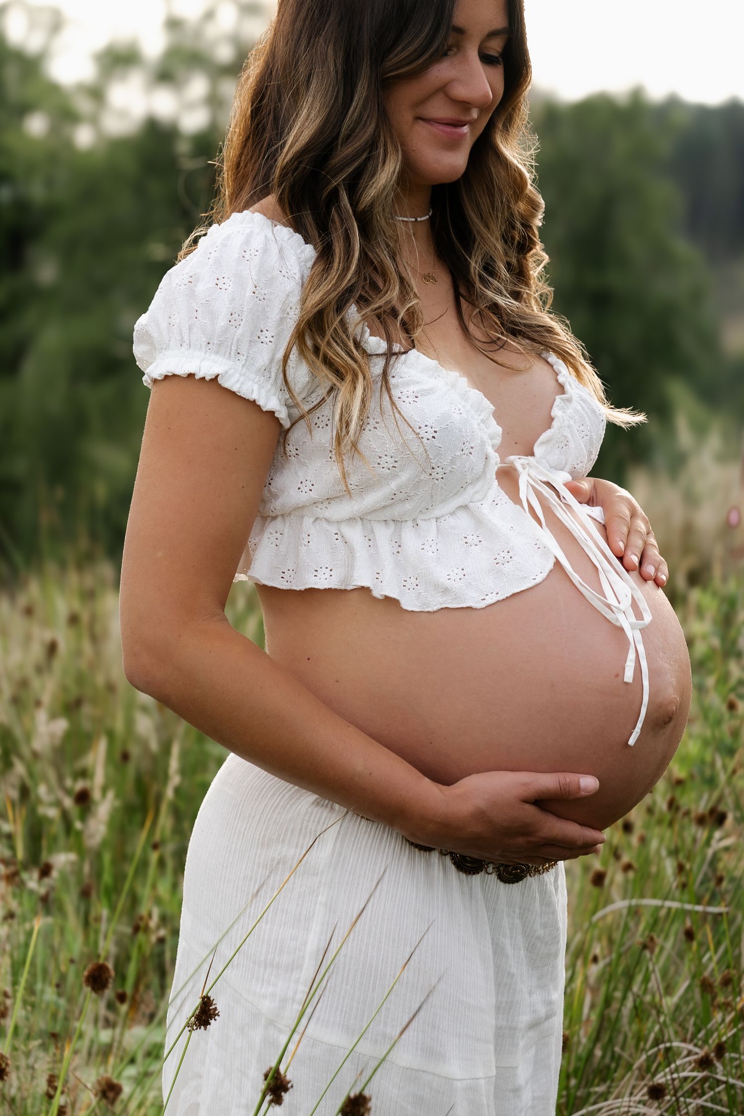 Smiling pregnant woman in white eyelet dress cradling baby bump in a meadow.