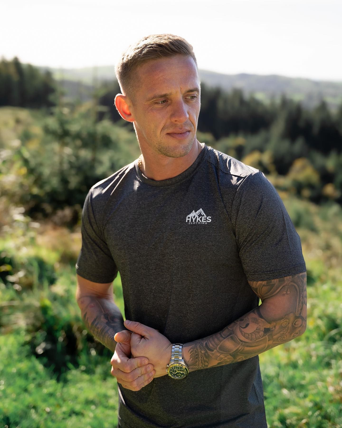 Tattooed man in dark gray Fitkes t-shirt and watch stands outdoors with green hills in background.