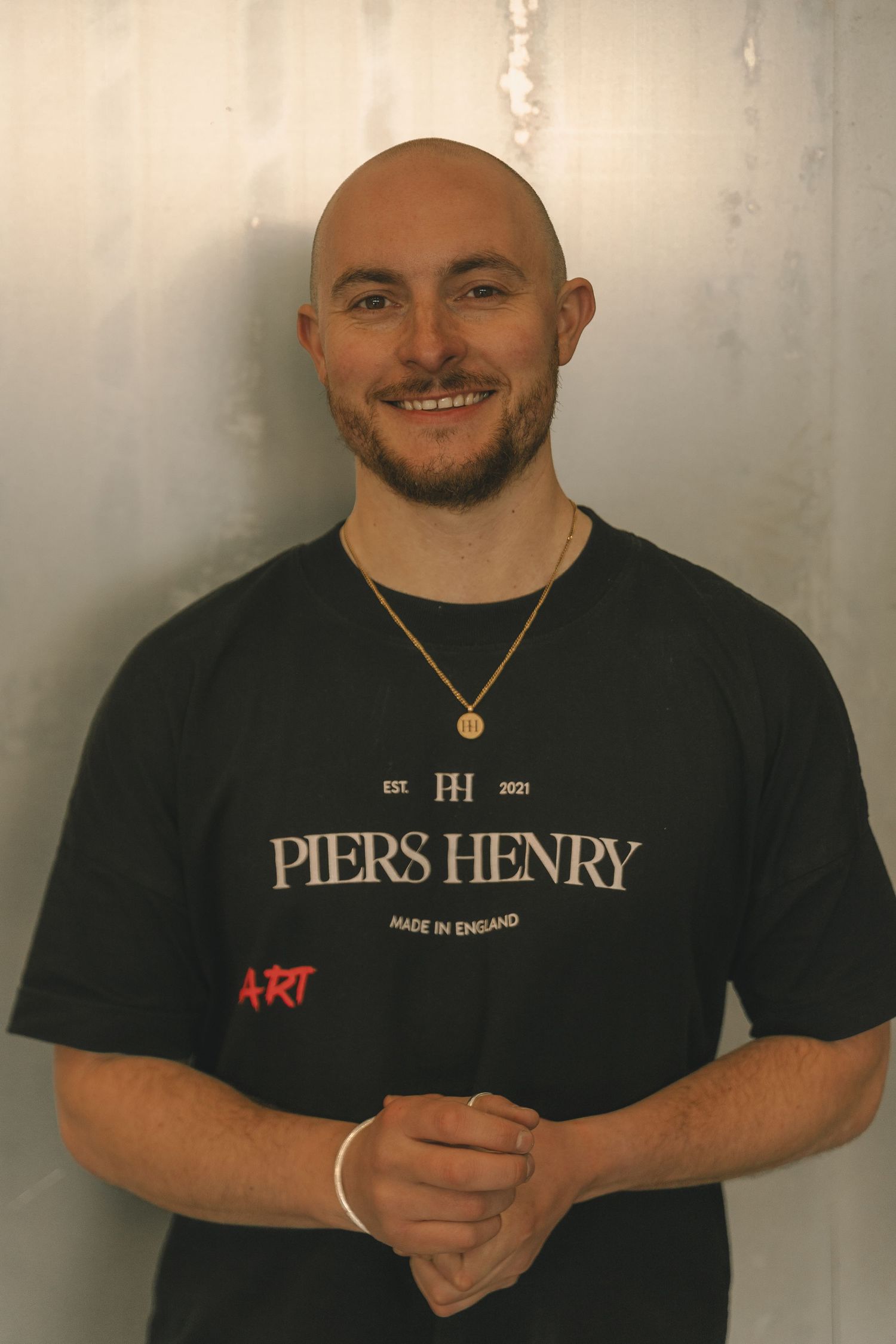 A smiling bald man wearing a black Piers Henry Made in England t-shirt and gold necklace poses indoors.