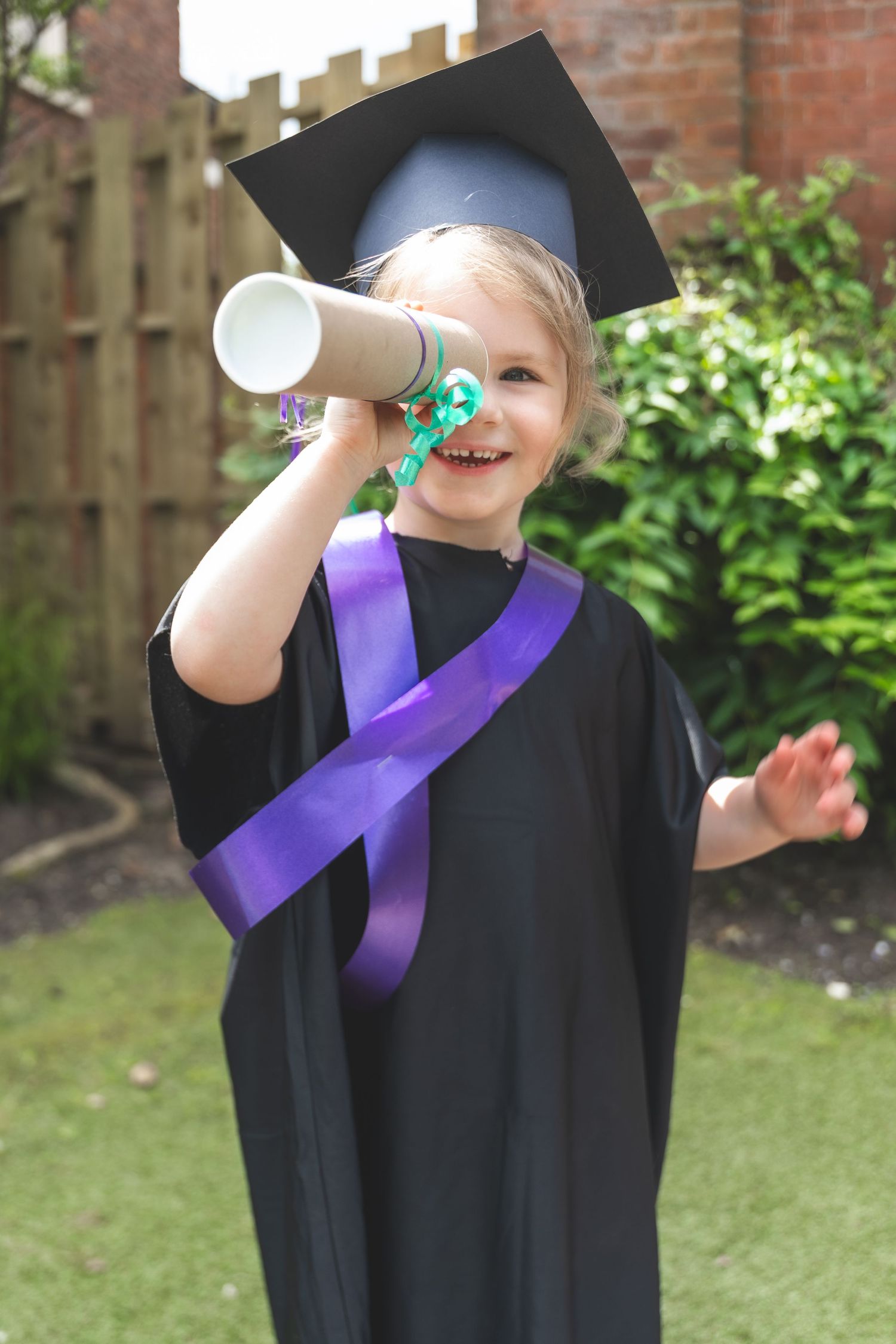 Young child in black graduation gown and cap holds diploma scroll, wearing purple sash, smiling outdoors.