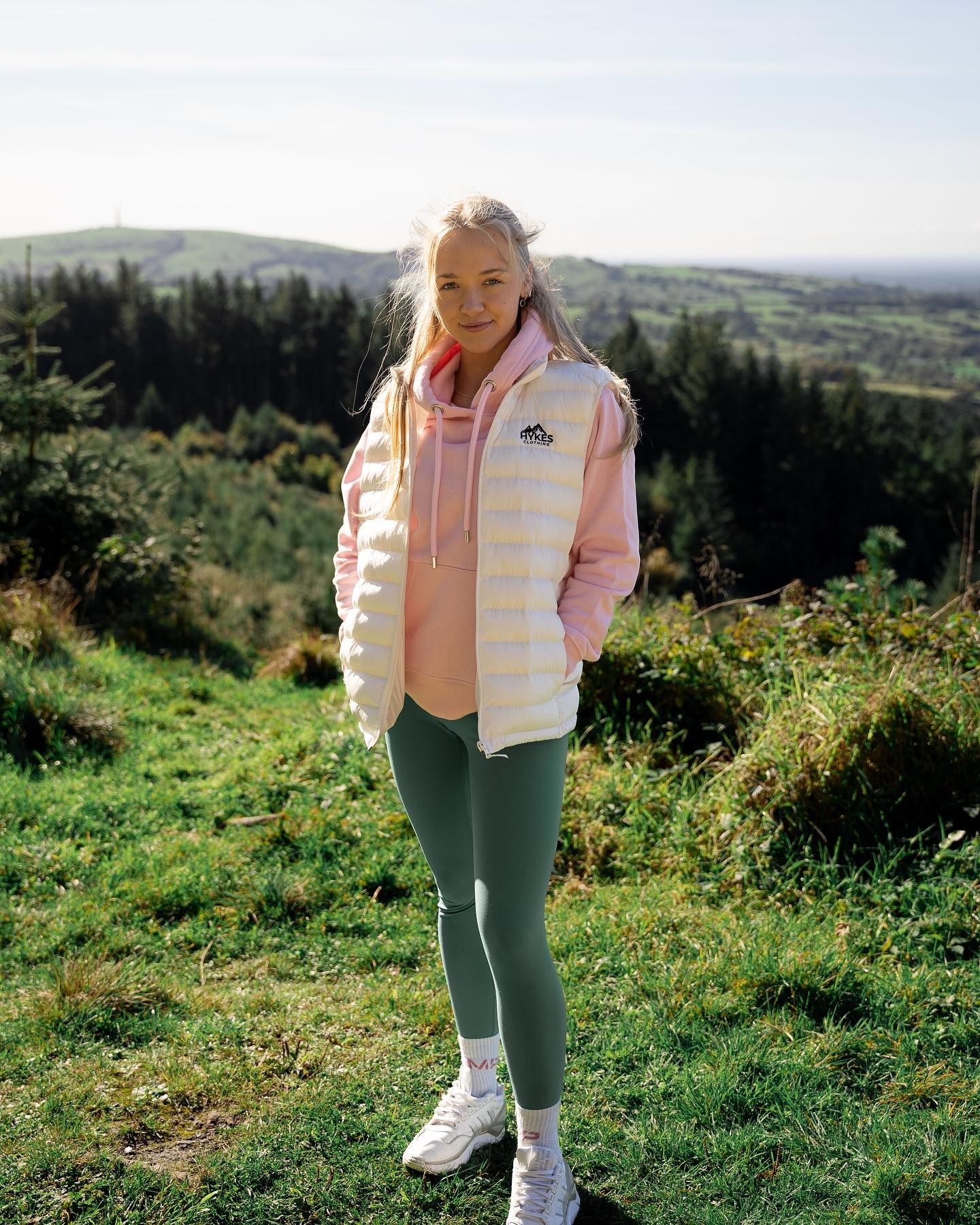 Young blonde woman in pink hoodie, white puffer vest, and green leggings standing on a hillside with scenic countryside views.