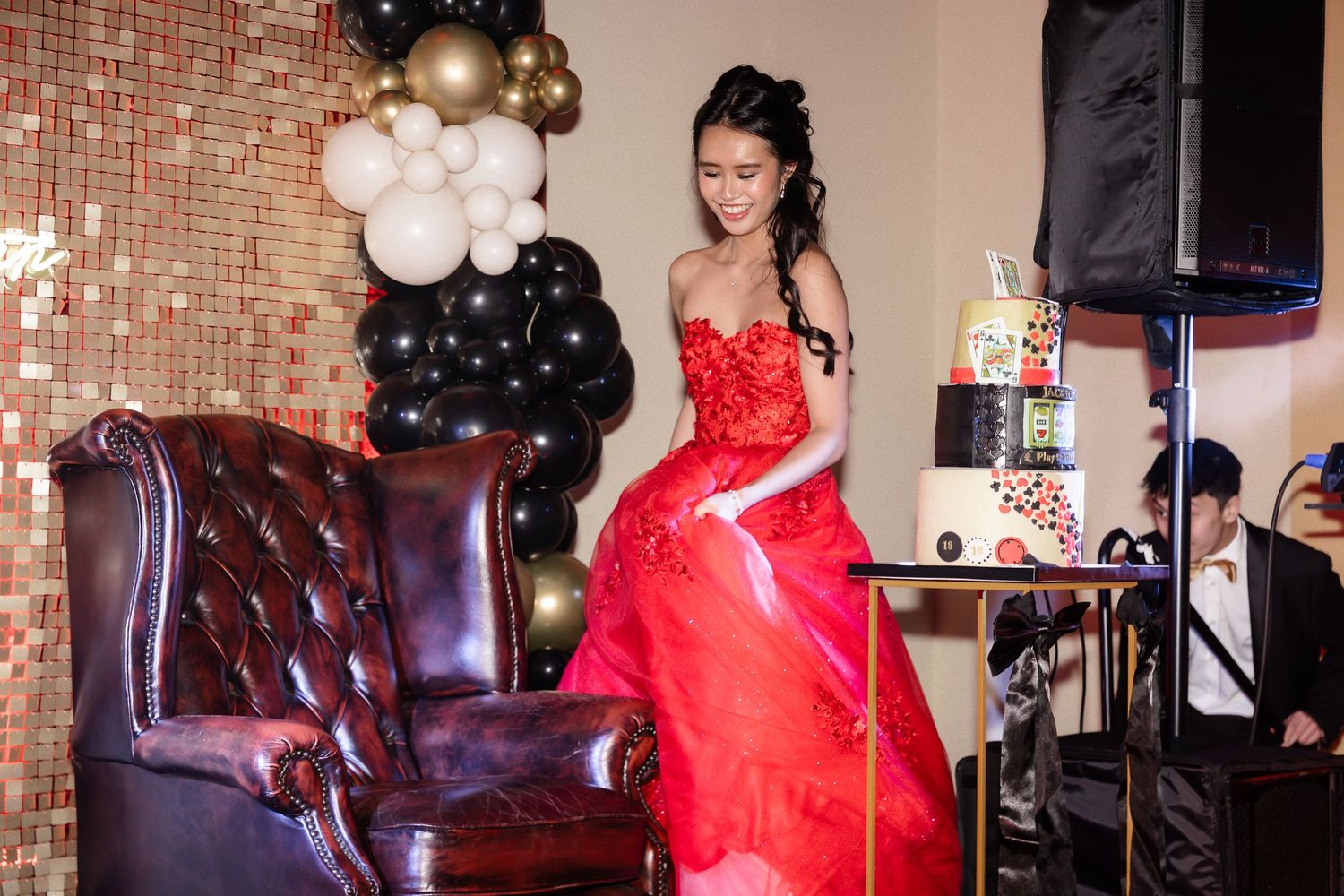 Young woman in elegant red ball gown poses near leather chair with celebration cake and balloons at formal party.
