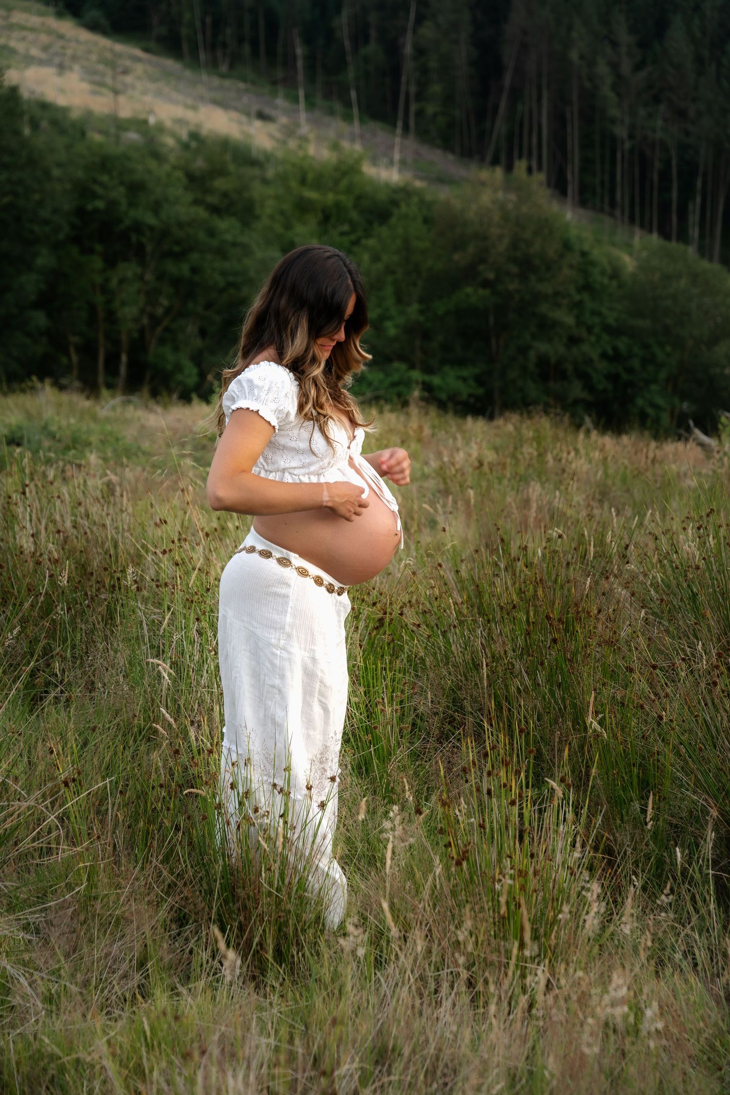 Pregnant woman in white outfit cradling her baby bump in a meadow.