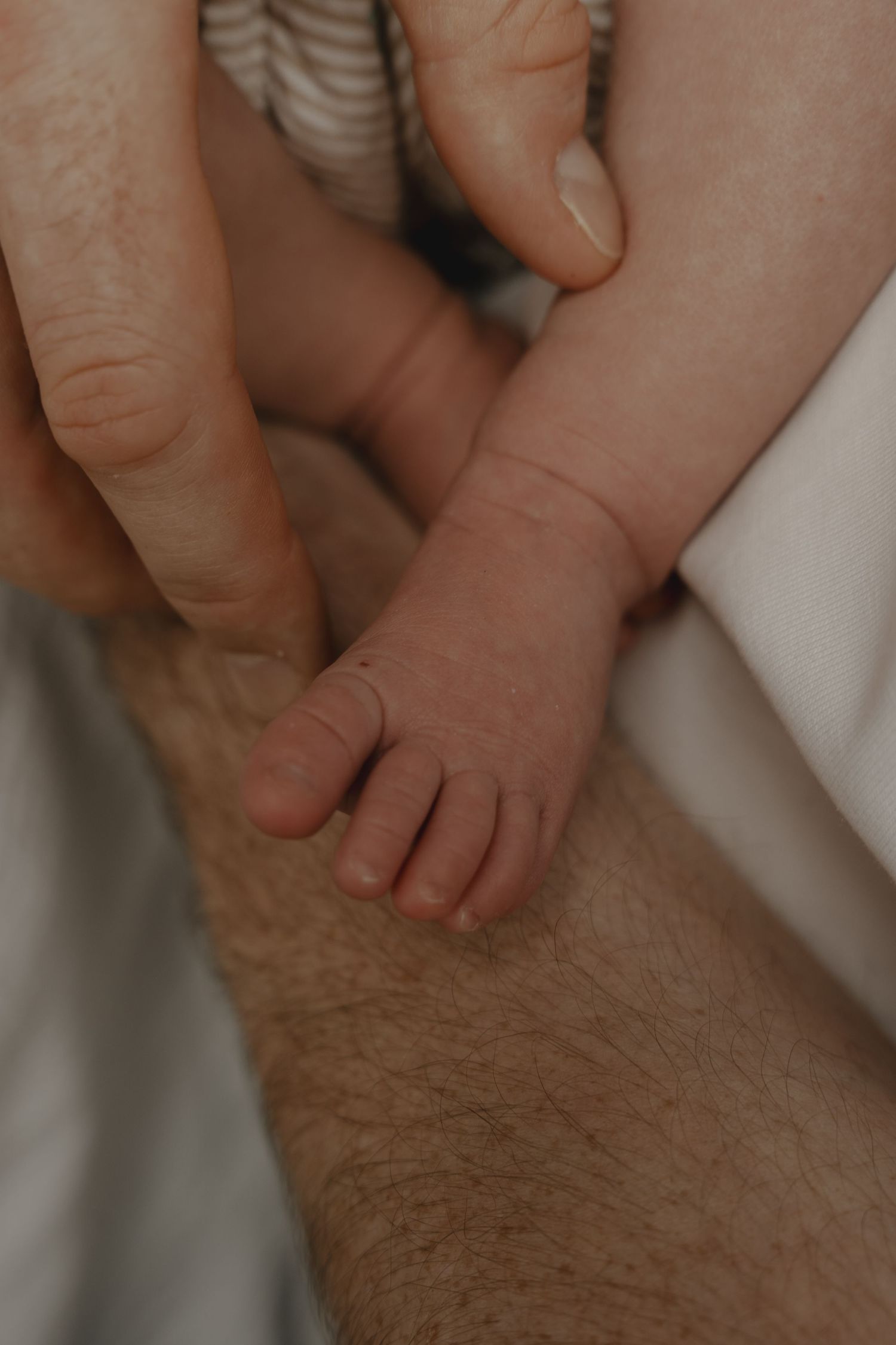 Tiny newborn baby foot resting on an adult's hairy leg, with a parent's hand gently cradling it.