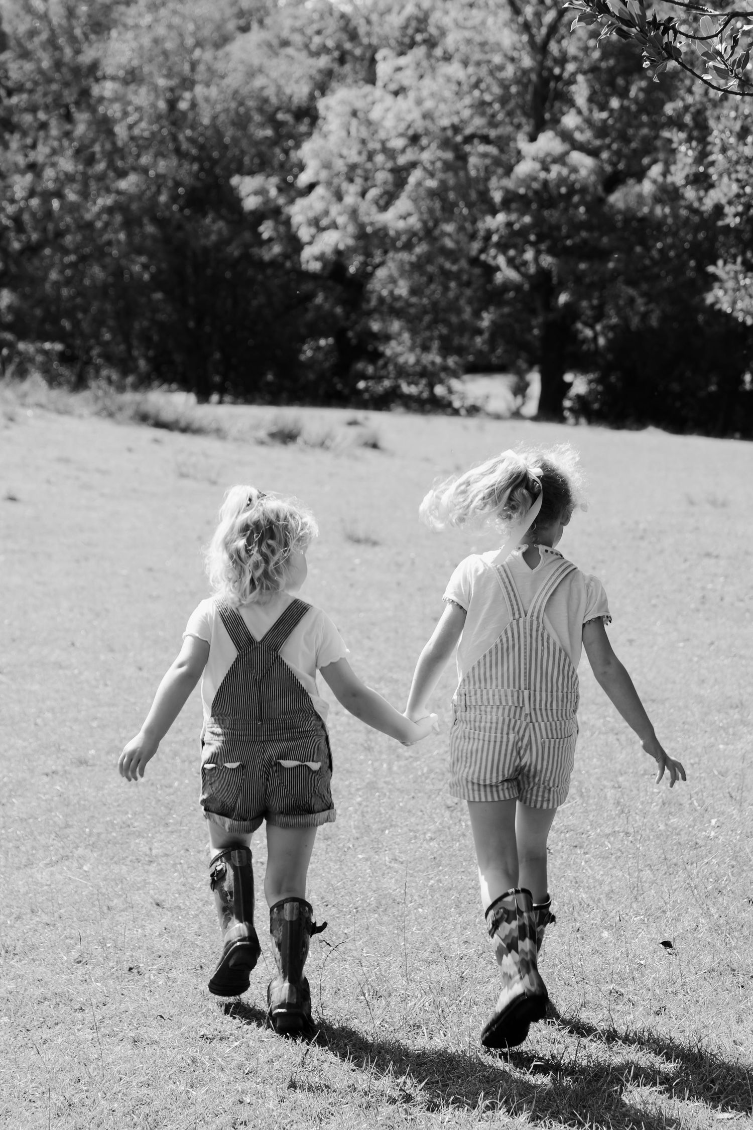 Black and white photo of two young girls holding hands walking in a sunny field.