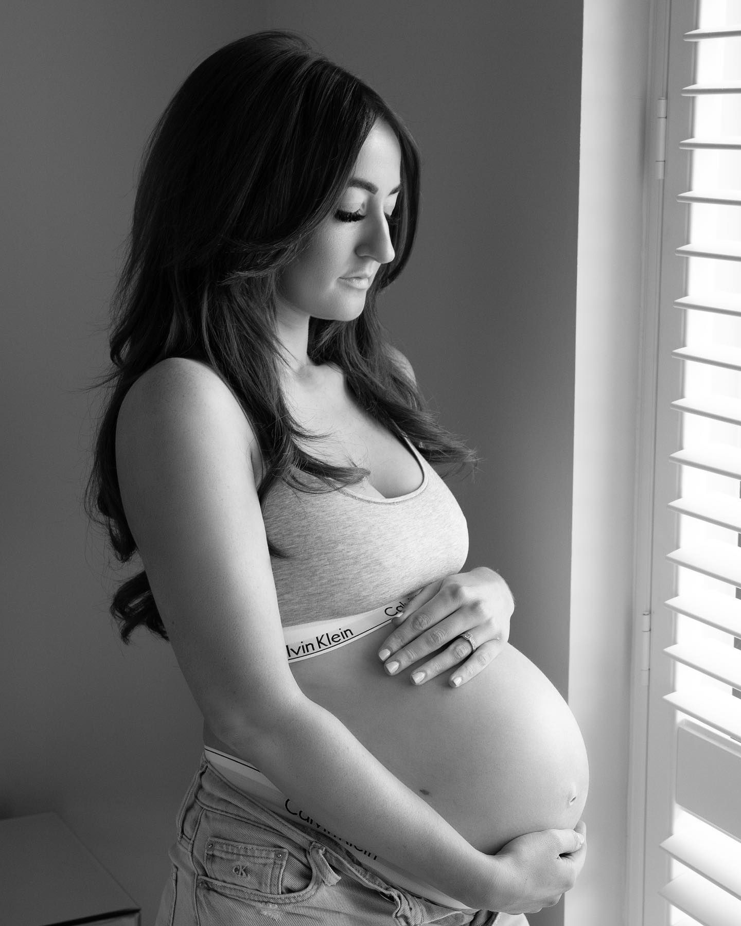 Black and white maternity portrait of pregnant woman holding baby bump near window.