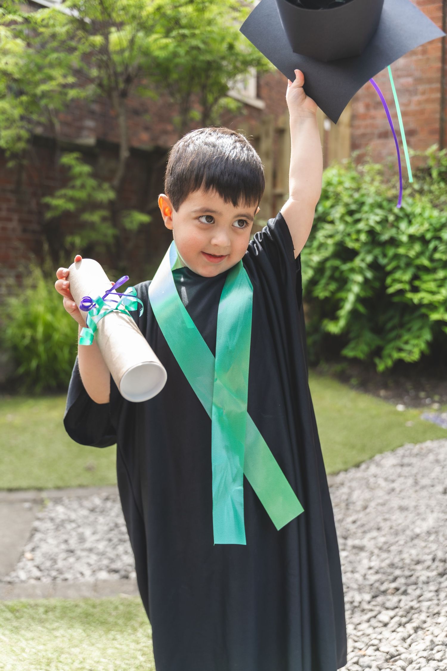 Young boy in black graduation gown and cap holds diploma outdoors, celebrating preschool graduation.