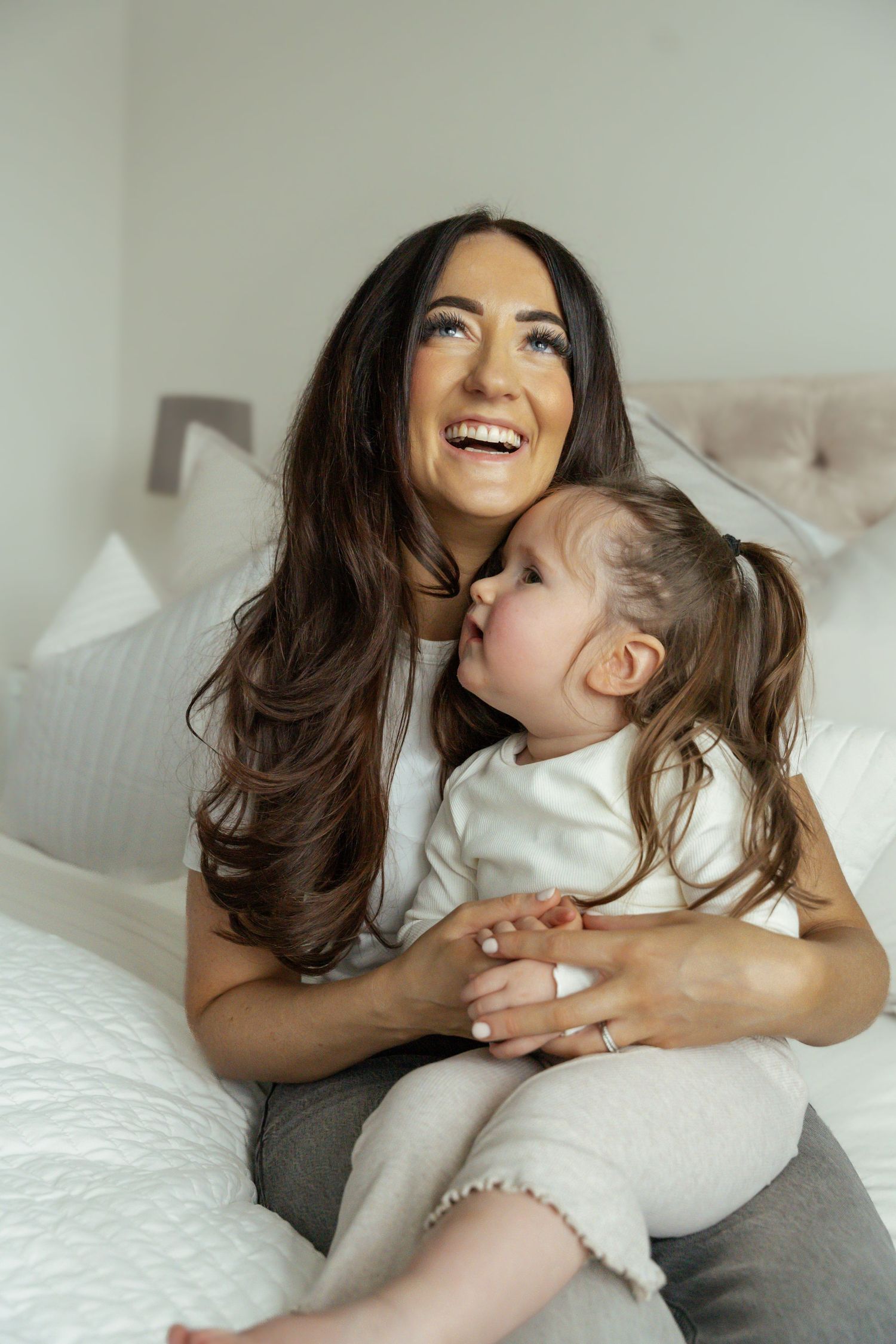 Laughing brunette mother cuddling young daughter on white bed in bright cozy bedroom.