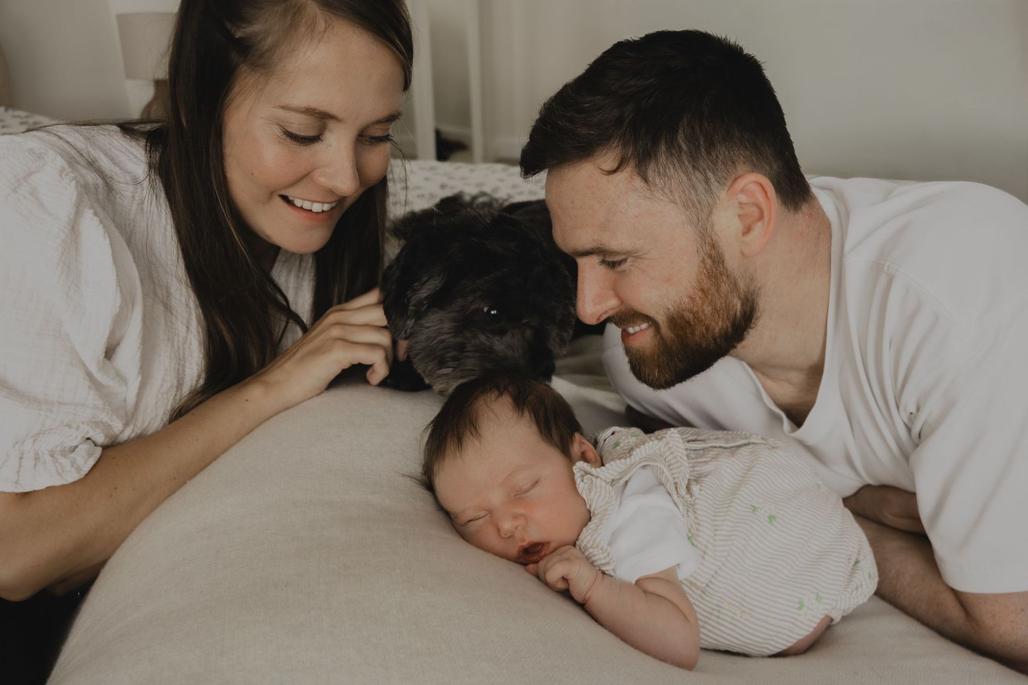 A couple smiles at their sleeping newborn baby on a bed, with a black dog looking on in a cozy home setting.