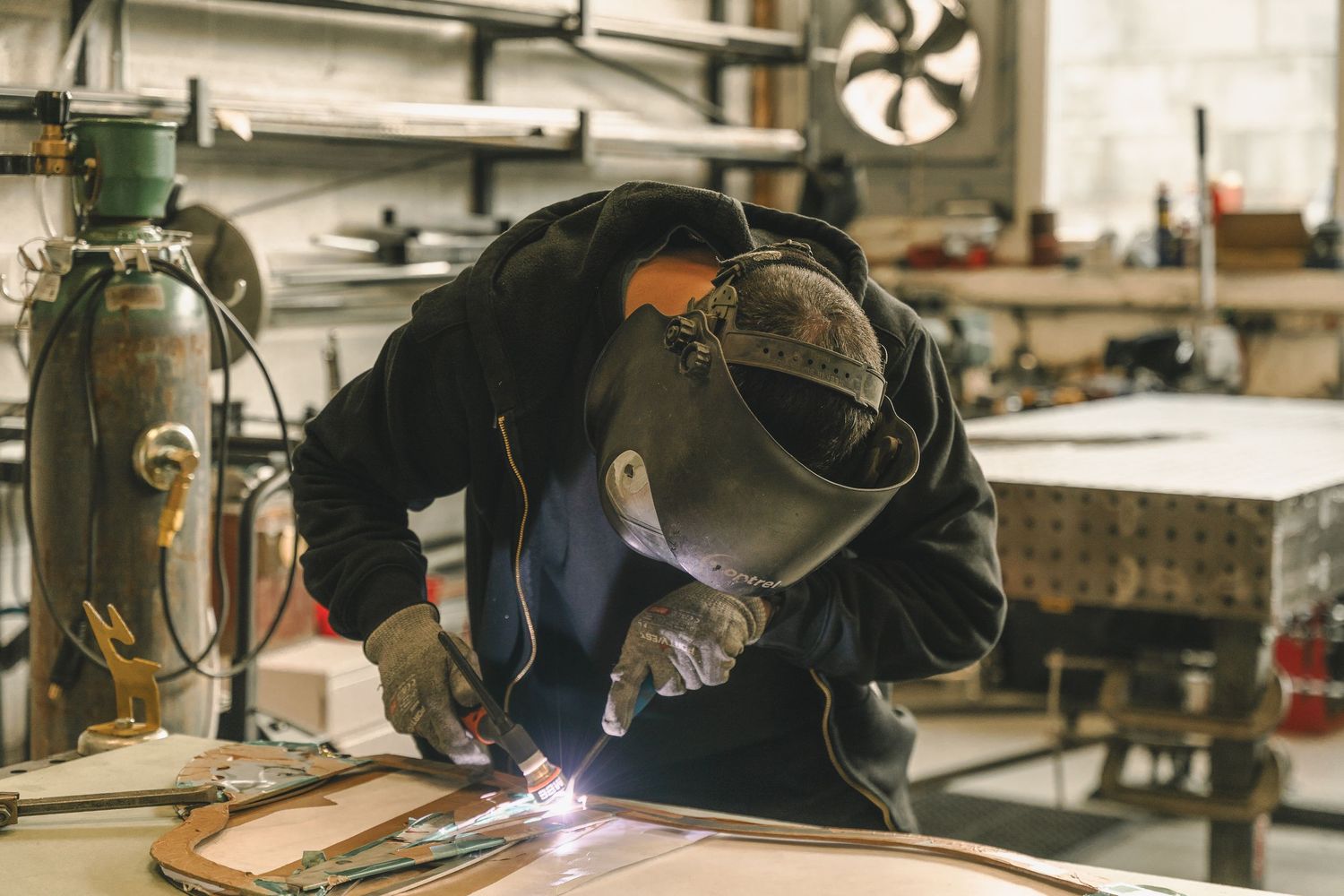 A welder in protective mask and gloves works on metal in an industrial workshop with bright welding sparks.