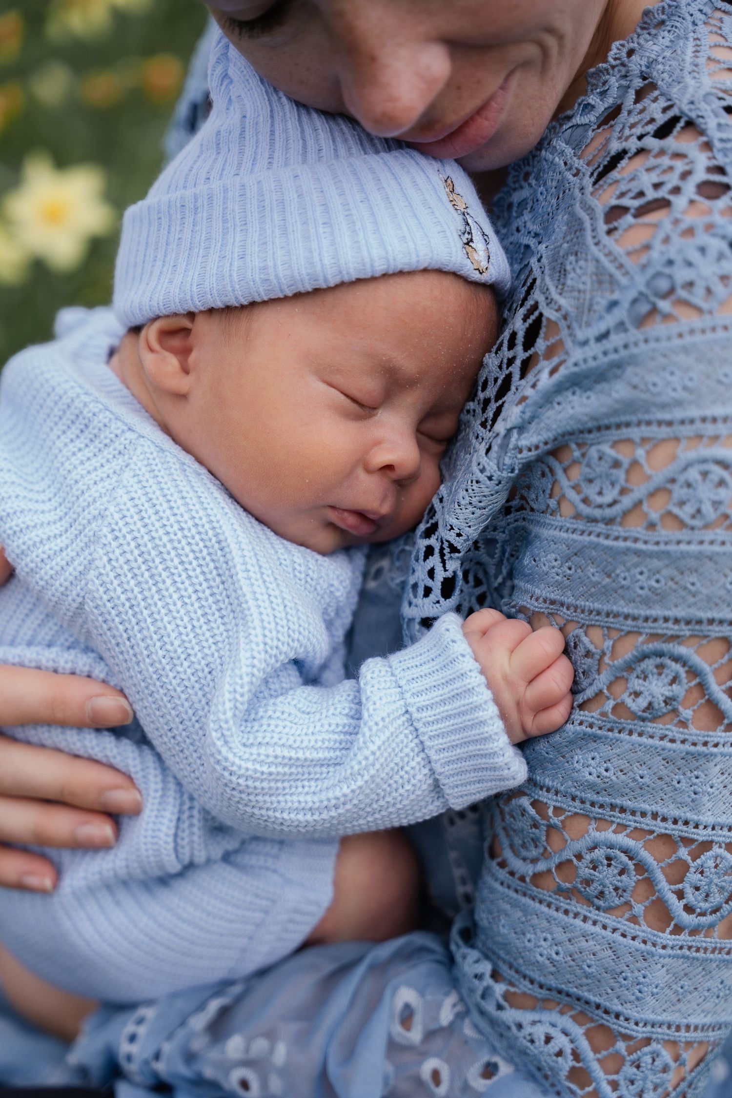 Mother tenderly cradling sleeping newborn in blue knit outfit against her chest outdoors.