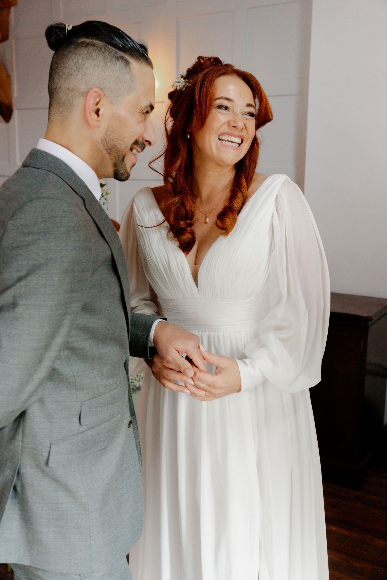 Smiling redhead bride in white gown exchanging rings with groom in grey suit at indoor wedding ceremony.
