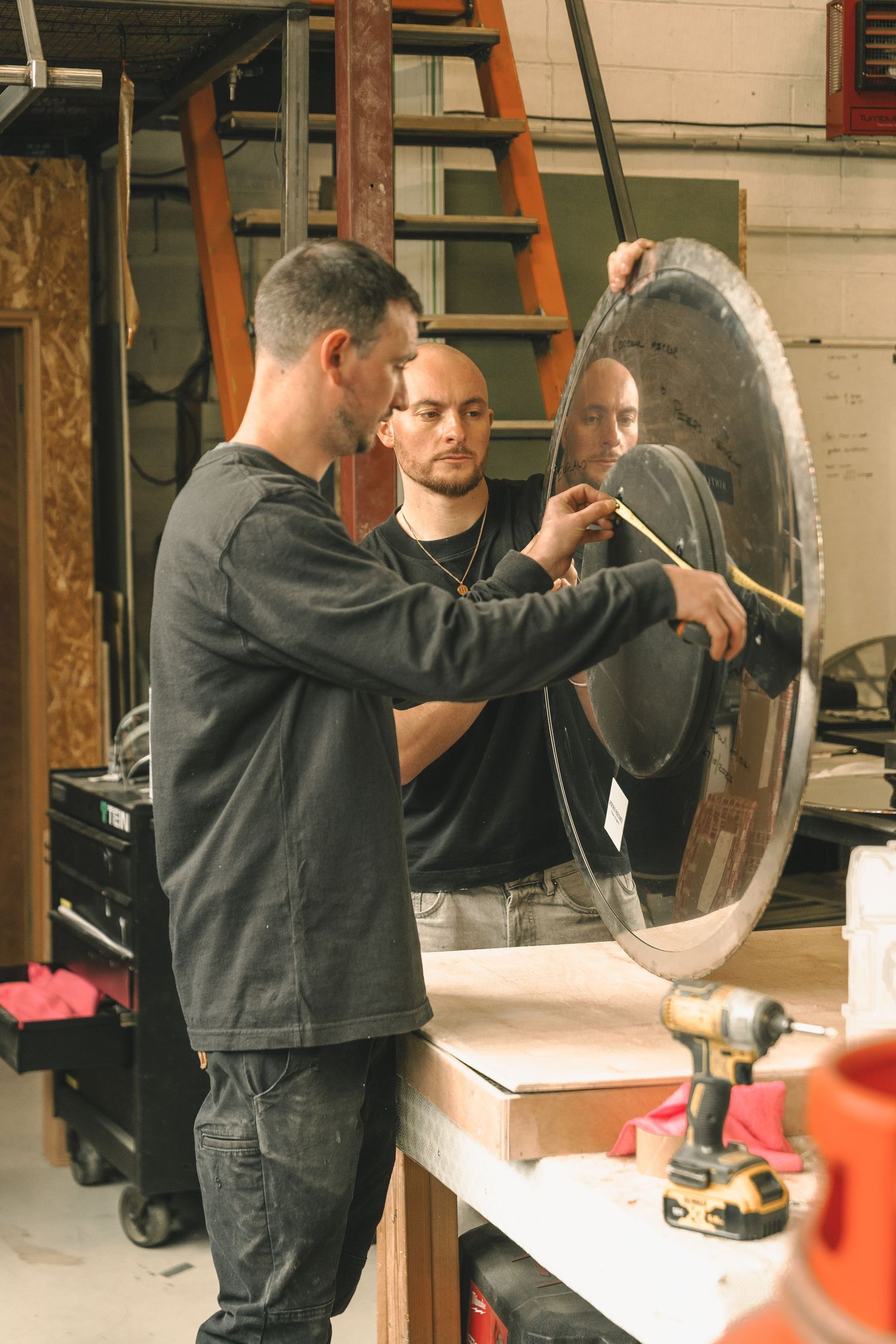 Two men examine and work on a large circular polished mirror mounted on a workbench in a workshop.