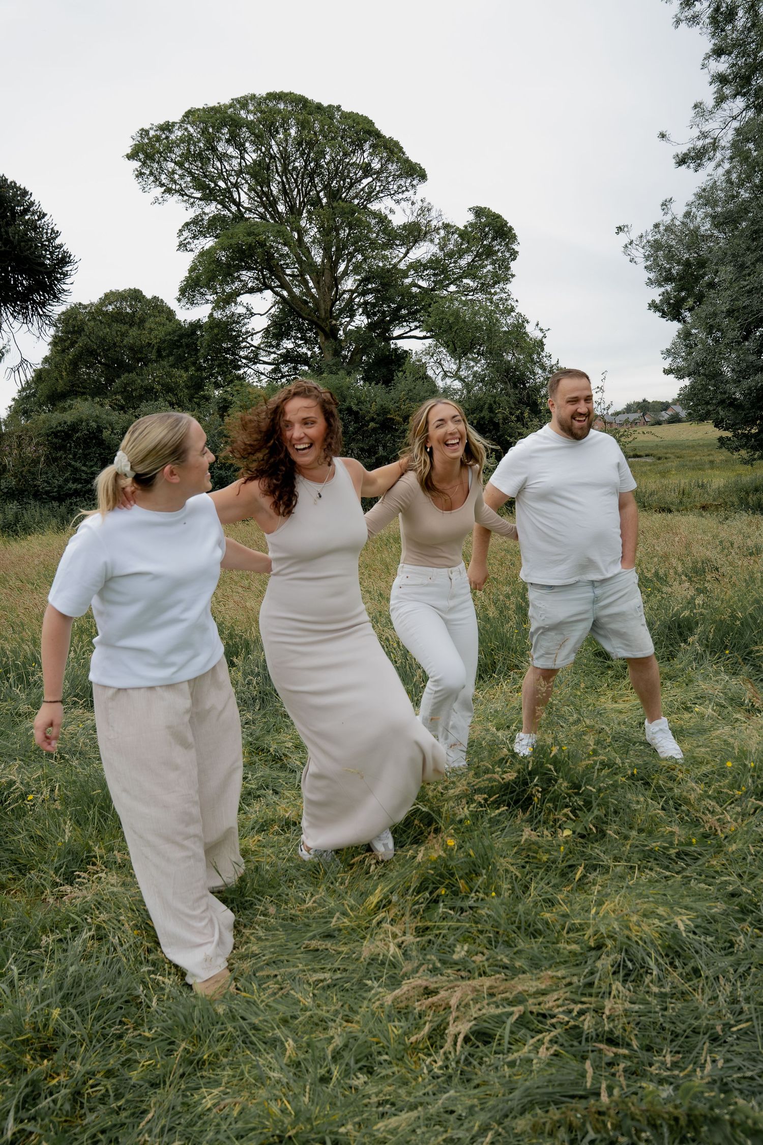 Four adults laughing and running together through green grass field on cloudy day.
