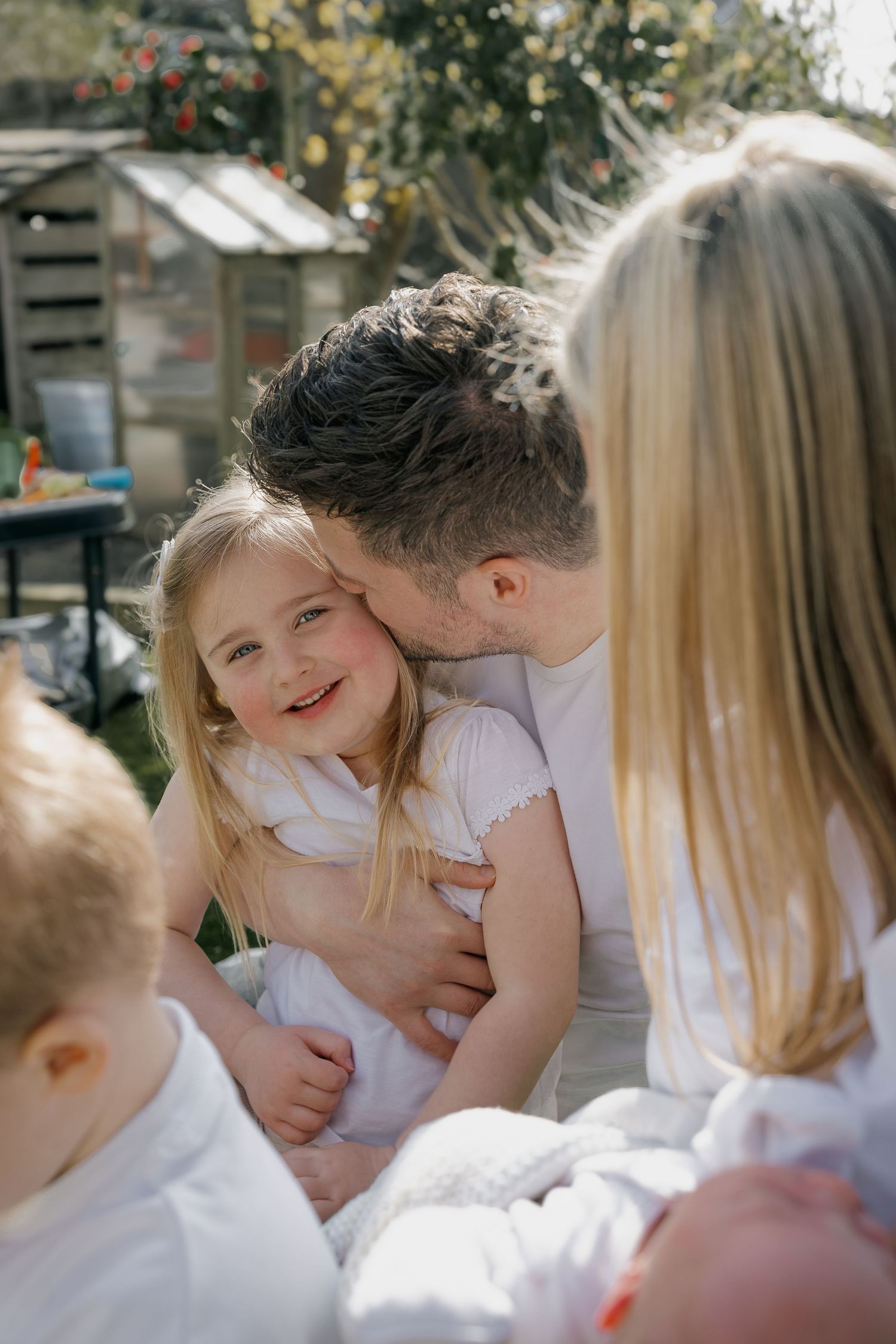 Father kissing his smiling blonde daughter on the cheek while sitting together in a sunny backyard garden.