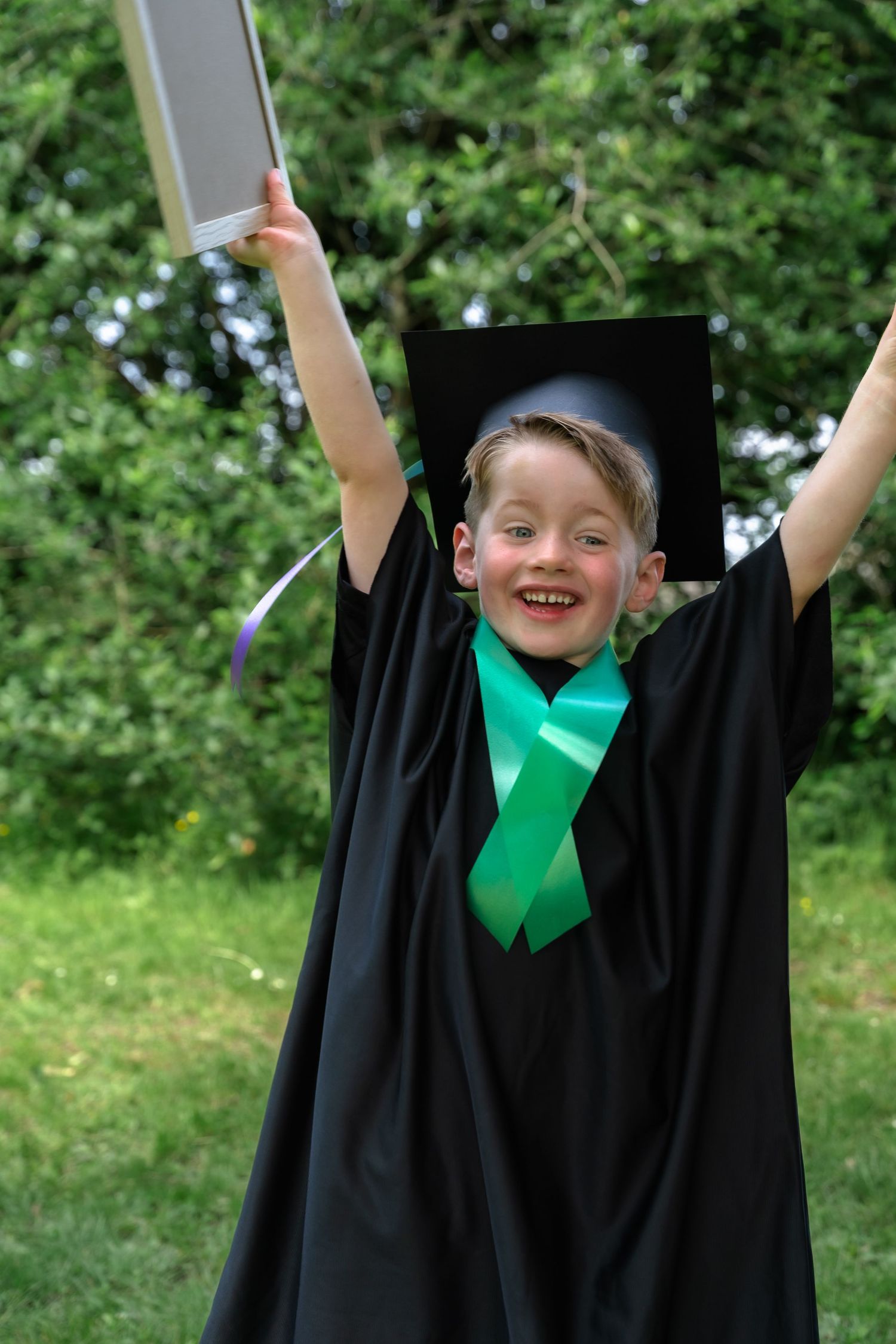 Young child in black graduation gown and cap raises diploma and arms in celebration outdoors.